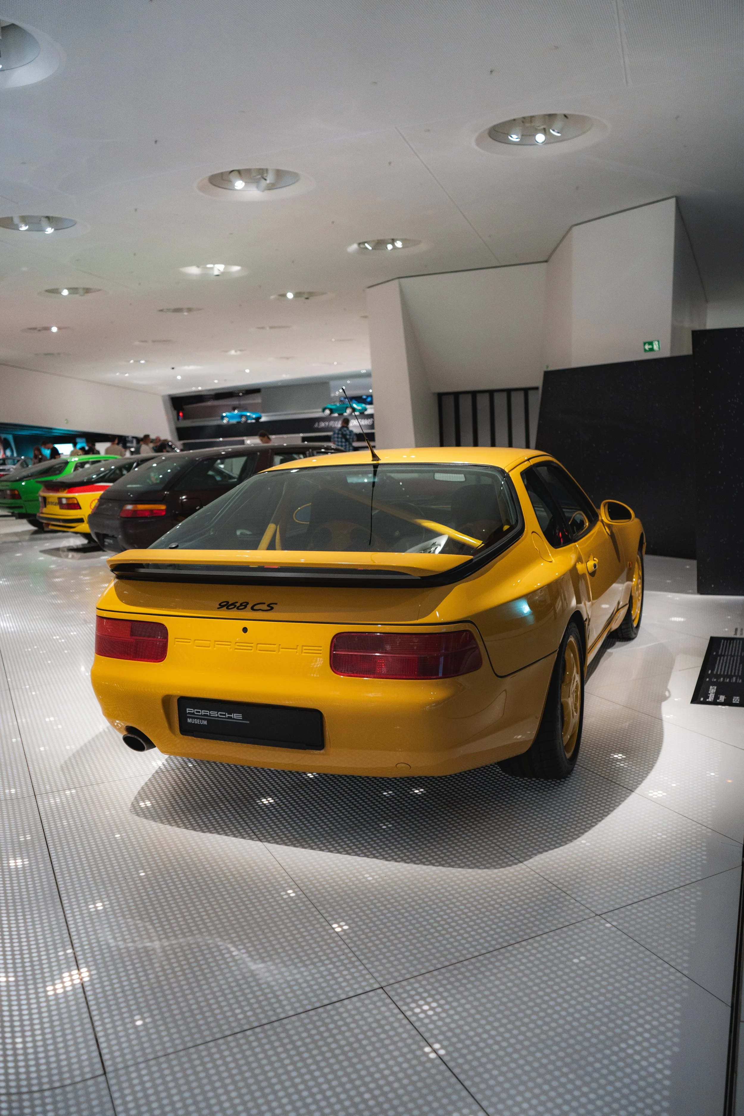 A yellow Porsche 968 CS on display in a museum, surrounded by other cars in the background.