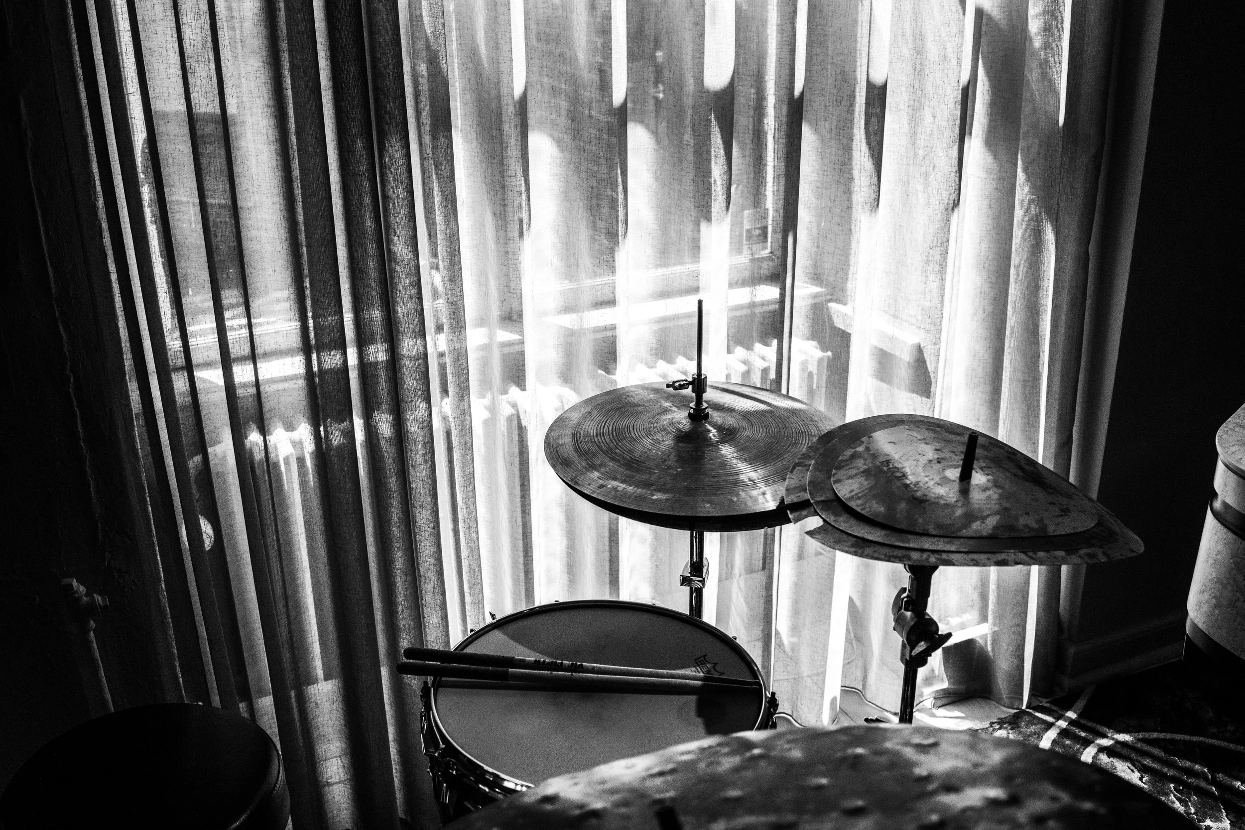 Black and white photo of a drum set near a window with sheer curtains, sunlight casting shadows on the drums.