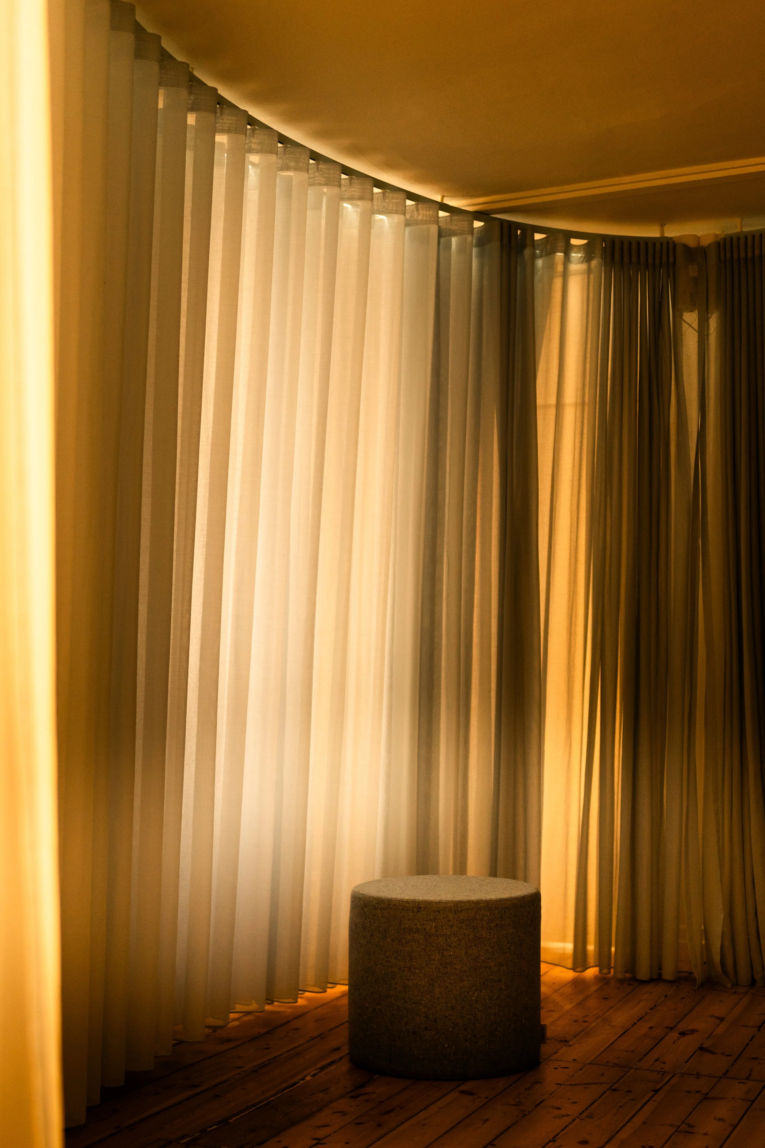 A room with multiple beige curtains hanging from the ceiling, with a round gray ottoman on a wooden floor.