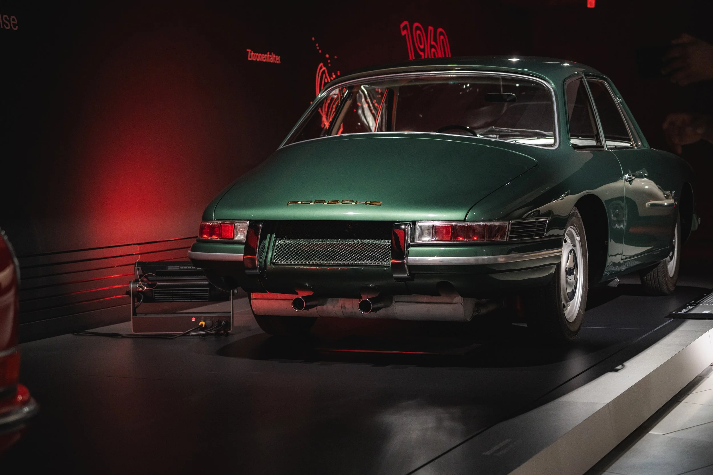 A classic green Porsche car displayed in a museum with a black background and red neon signs.