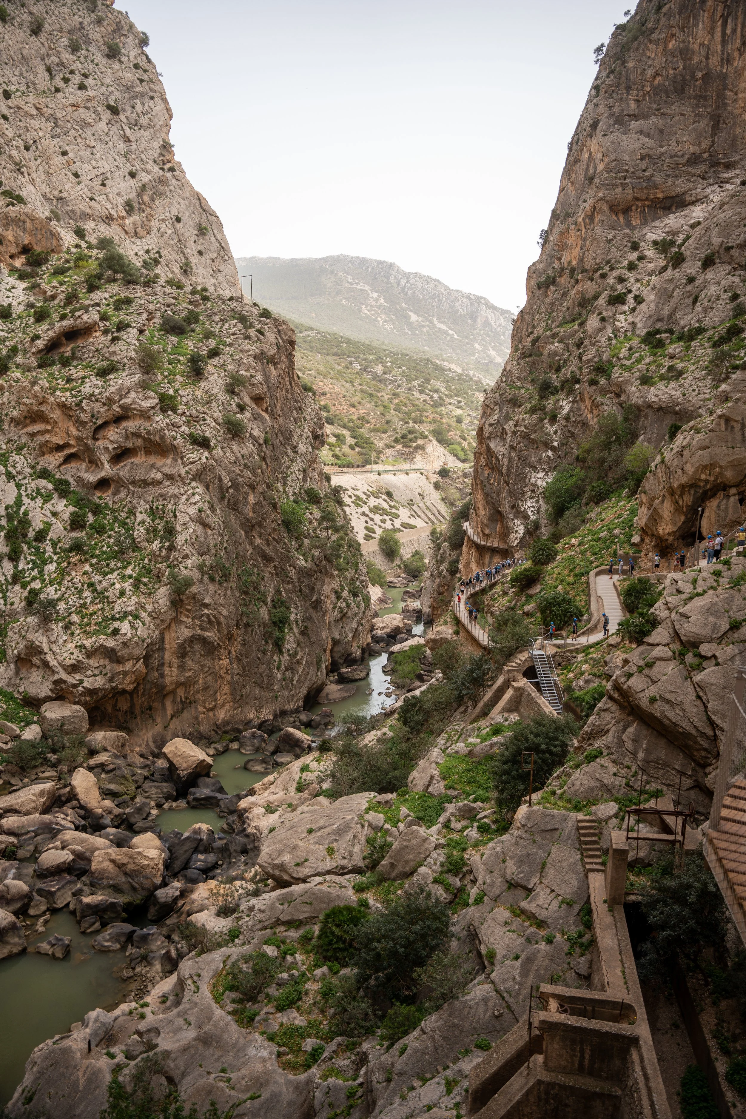 A narrow river flows through a deep canyon with rocky cliffs and a walkway carved into the rockside, where people are walking.