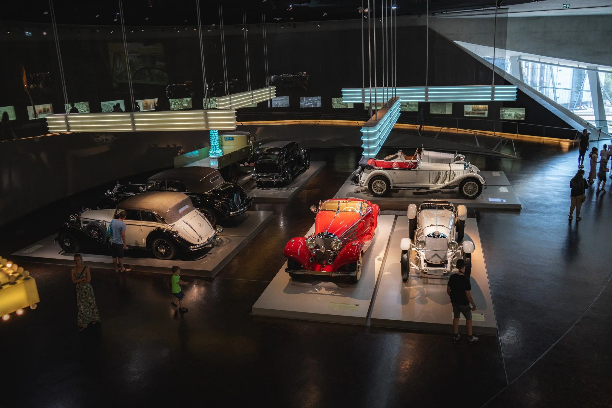 Several vintage cars displayed in a museum or exhibit, illuminated by overhead lighting, with visitors viewing the cars.