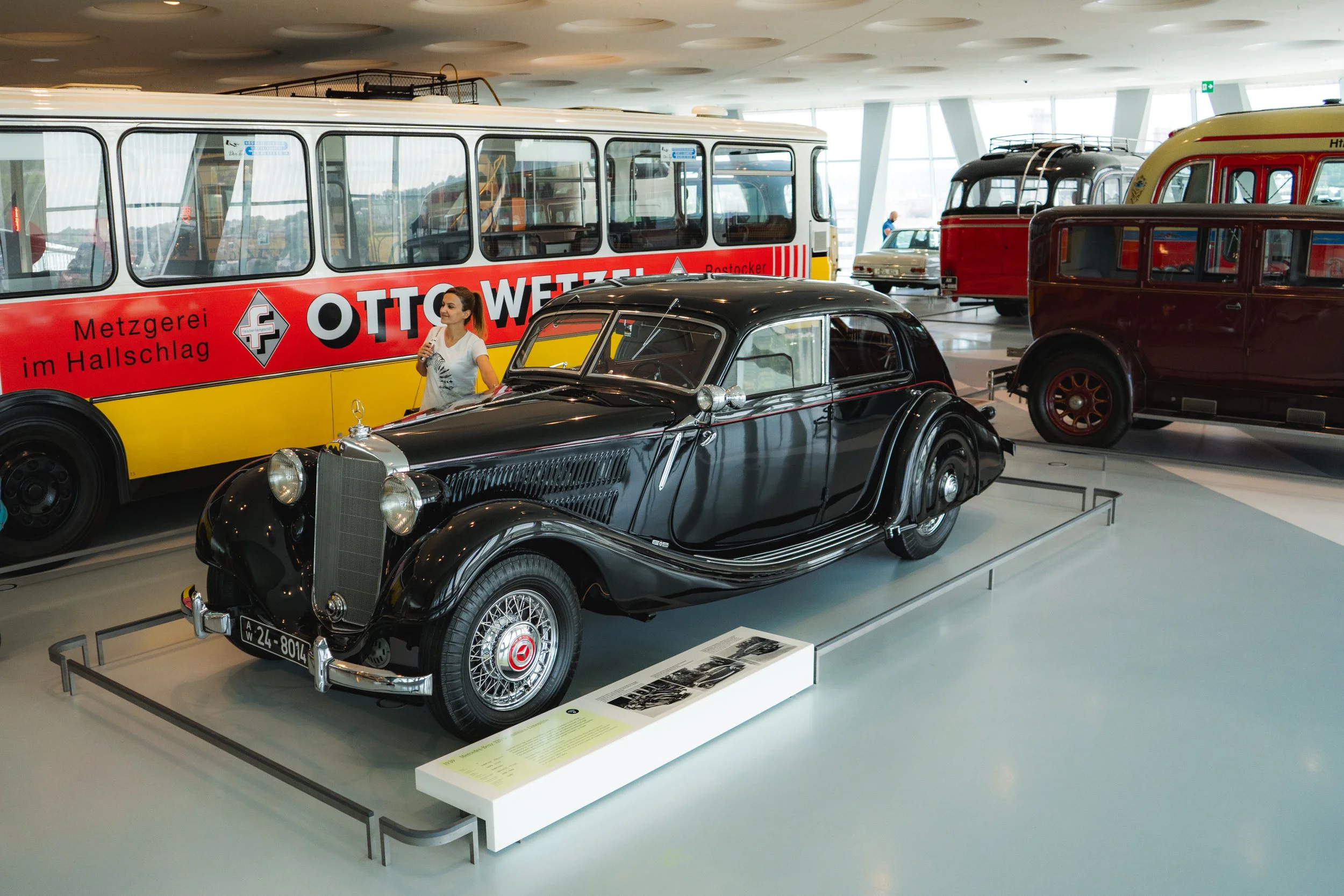 A black vintage car displayed in a museum with other vintage vehicles and a yellow bus with red and white accents in the background.