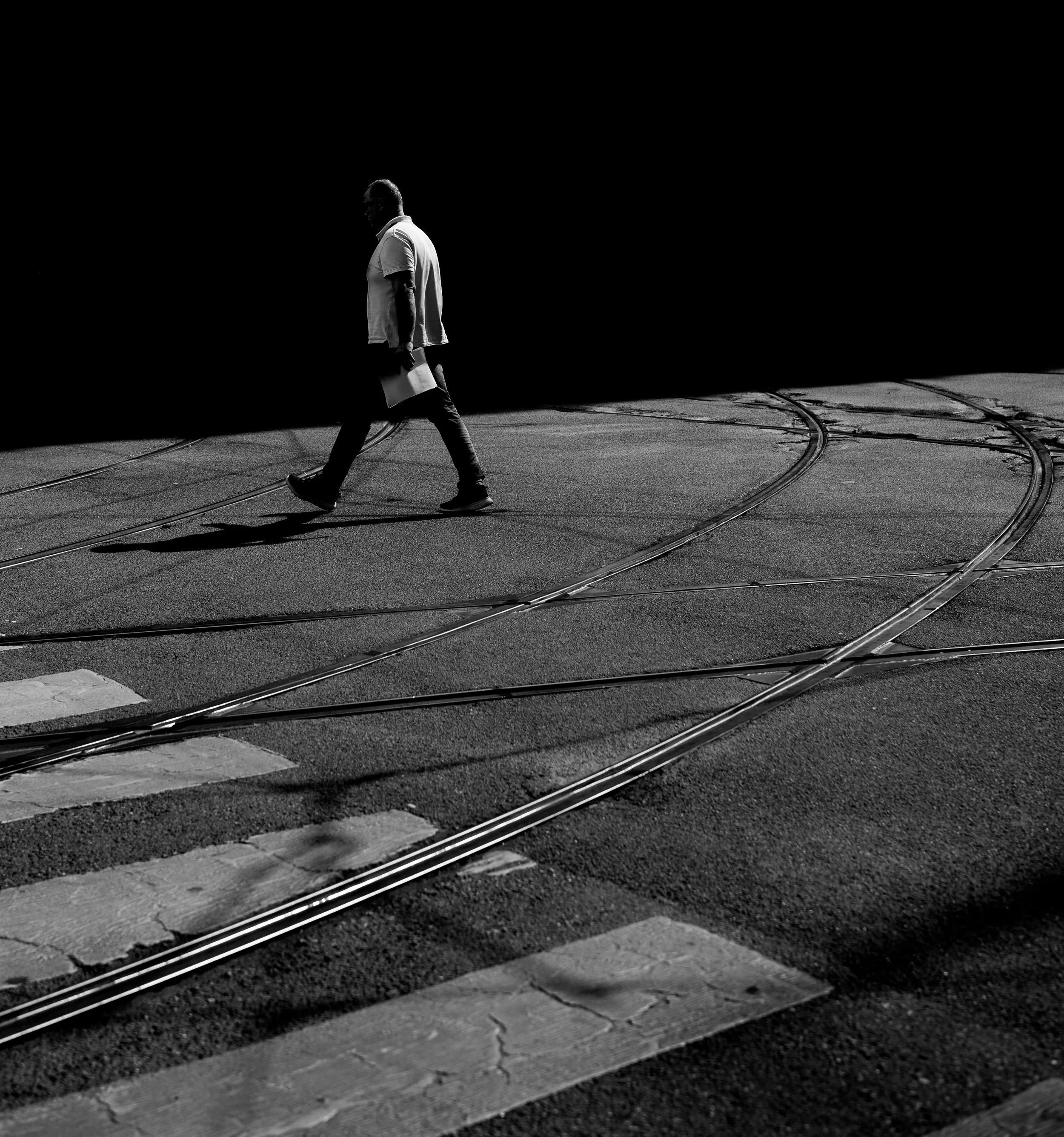 A man walking across a street with tram tracks at night in black and white, casting a shadow.