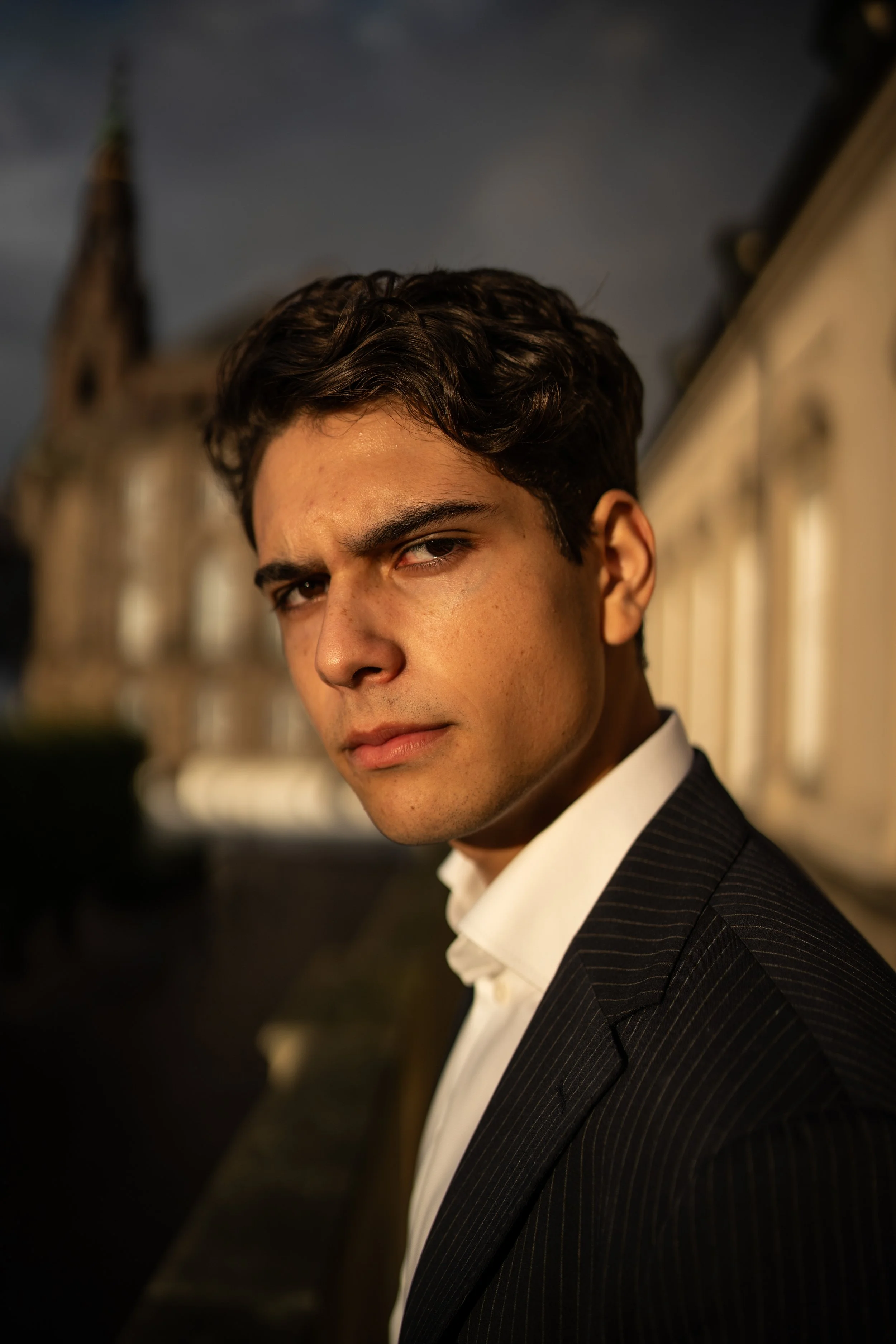 A young man with dark curly hair, wearing a black pinstripe suit and white shirt, looking serious while standing outdoors near a historic building during dusk.