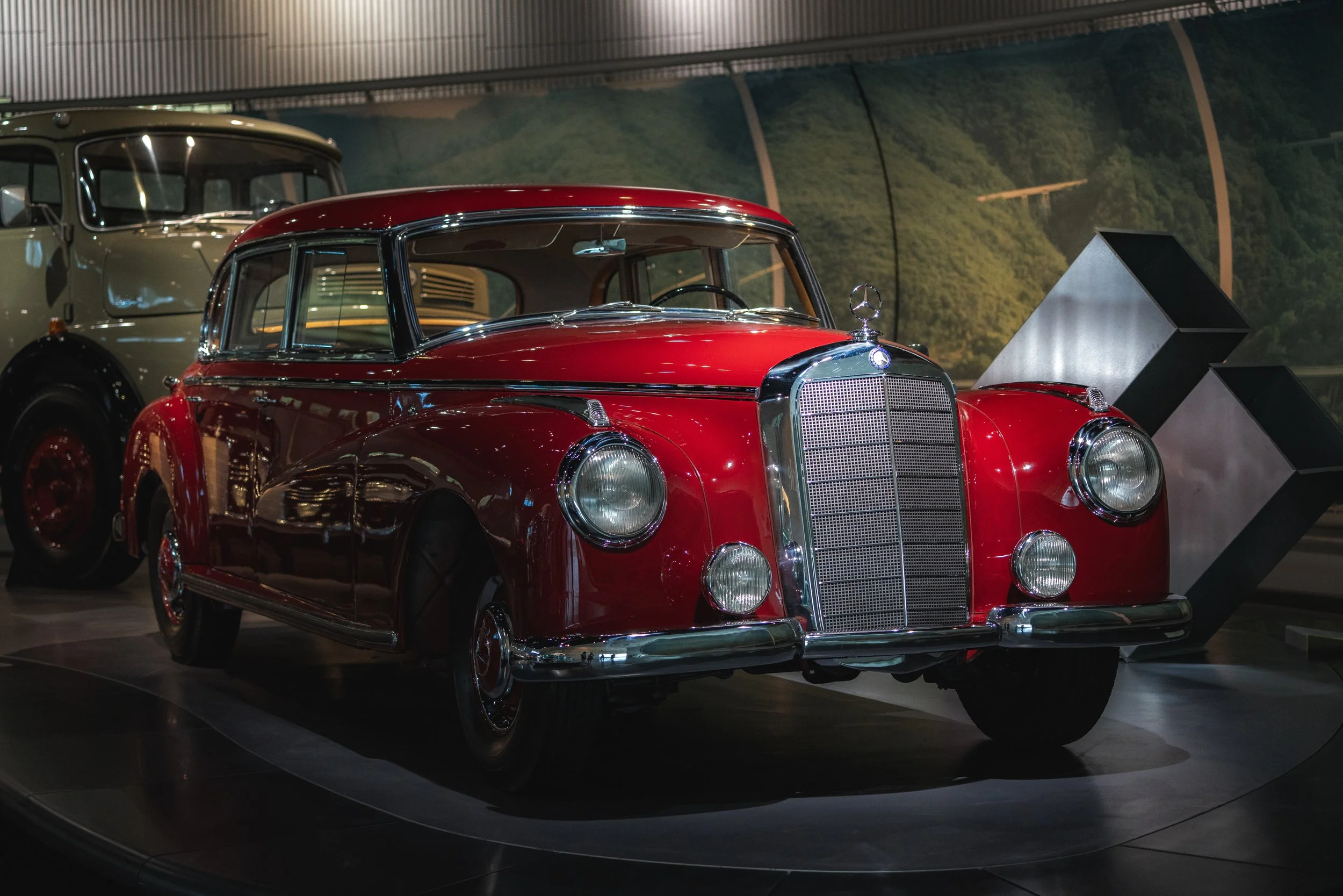 A shiny red vintage Mercedes-Benz automobile on display in a museum, with another classic vehicle in the background and a modern geometric sculpture nearby.