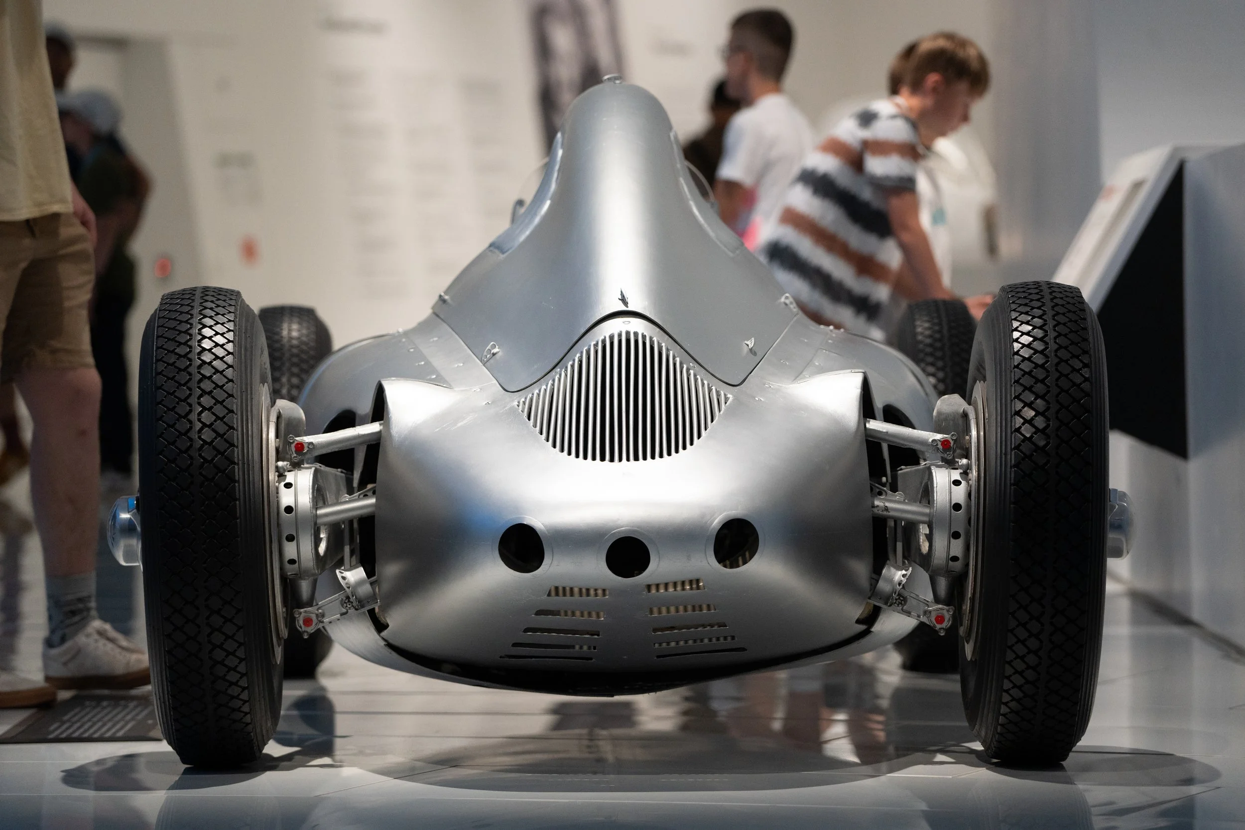 A vintage silver race car displayed at a museum with visitors observing in the background.