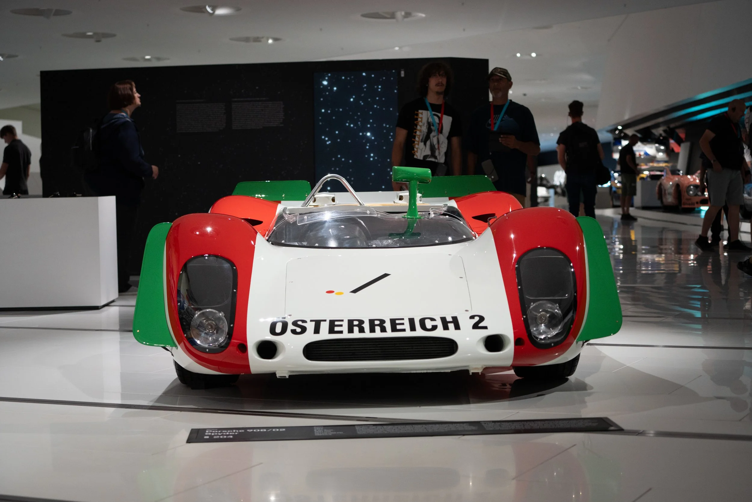 A vintage race car with a white, red, and green color scheme on display at a museum. The car has the word "Österreich 2" on the front and a sleek, aerodynamic design with open cockpit.
