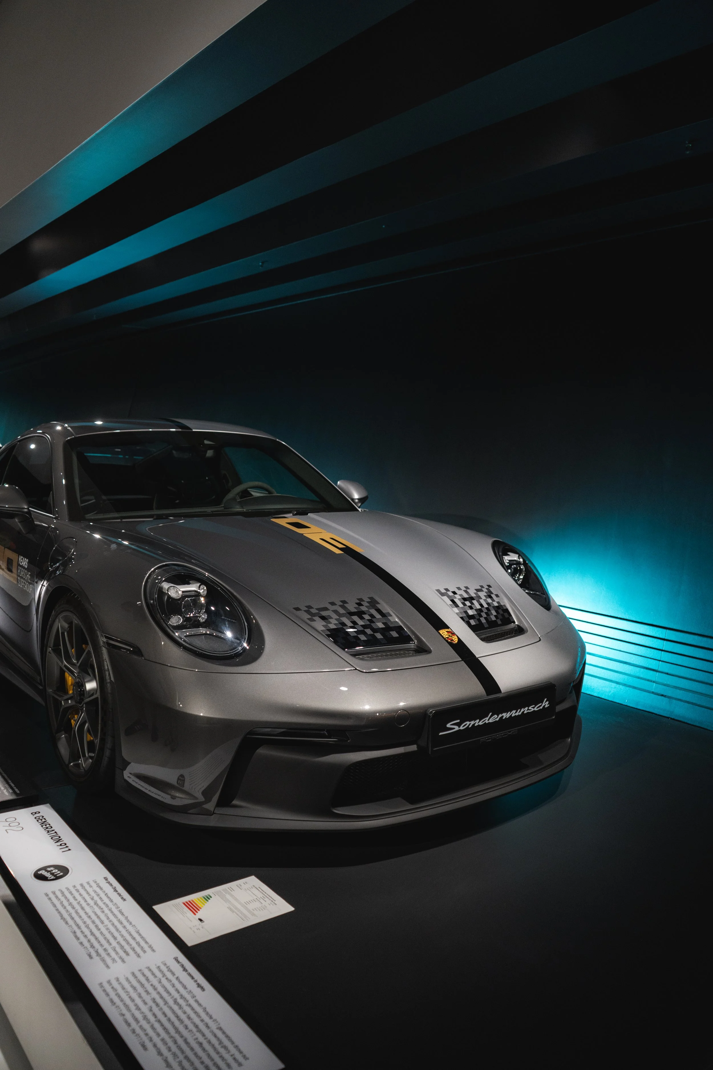 A silver Porsche racing car with black and gold decals on display at a car exhibit, illuminated by blue lights in a dimly lit space.