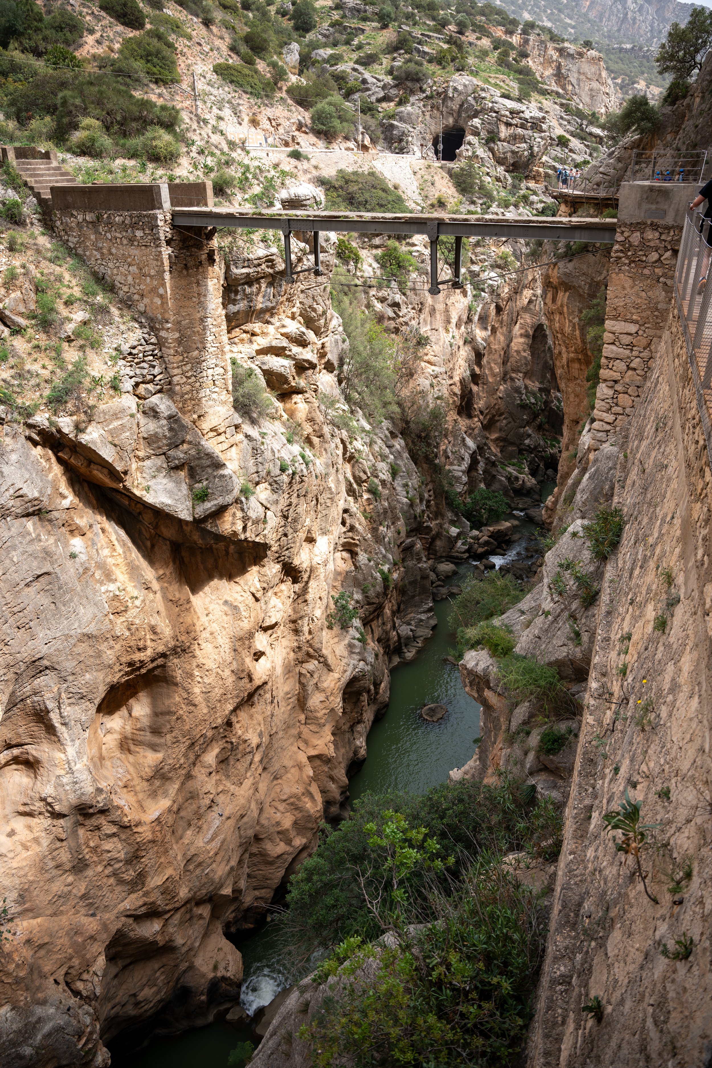 A narrow canyon with steep rock walls and a small river flowing through it. There is a bridge crossing over the canyon, and a tunnel in the hillside in the background.