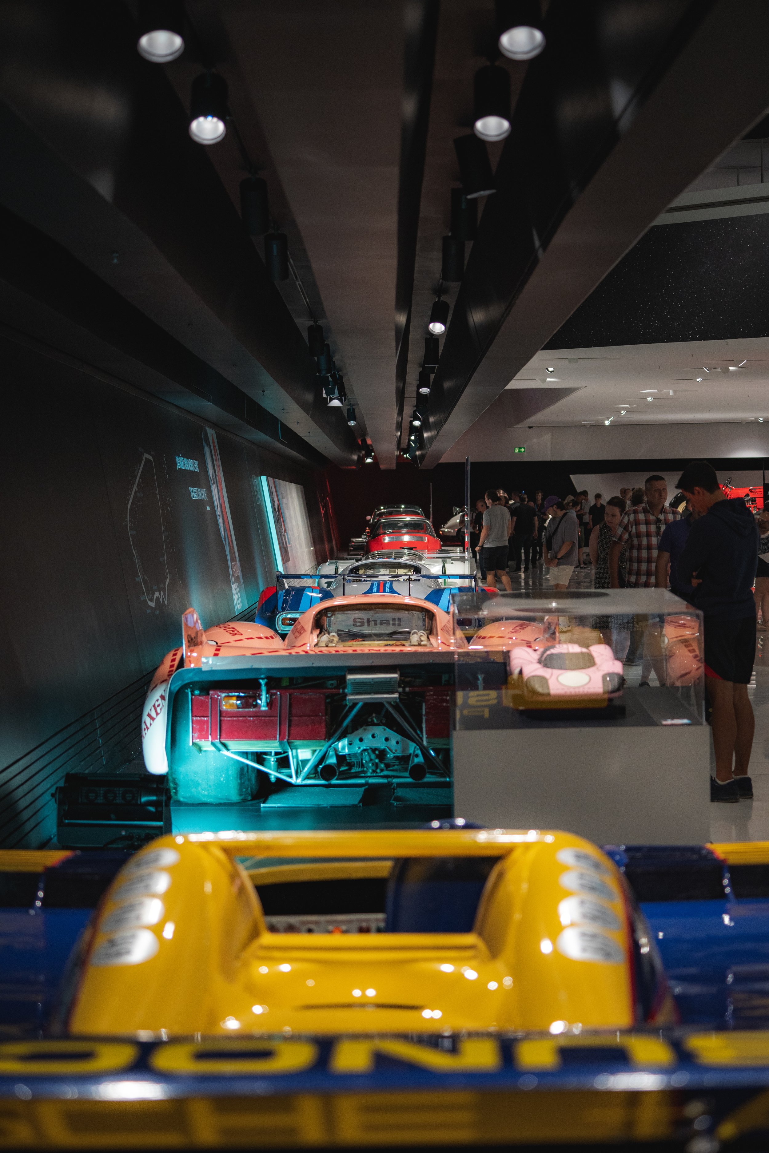 View of a car exhibition with race cars displayed indoors, people observing the cars, and a dark interior with spotlights on the ceiling.
