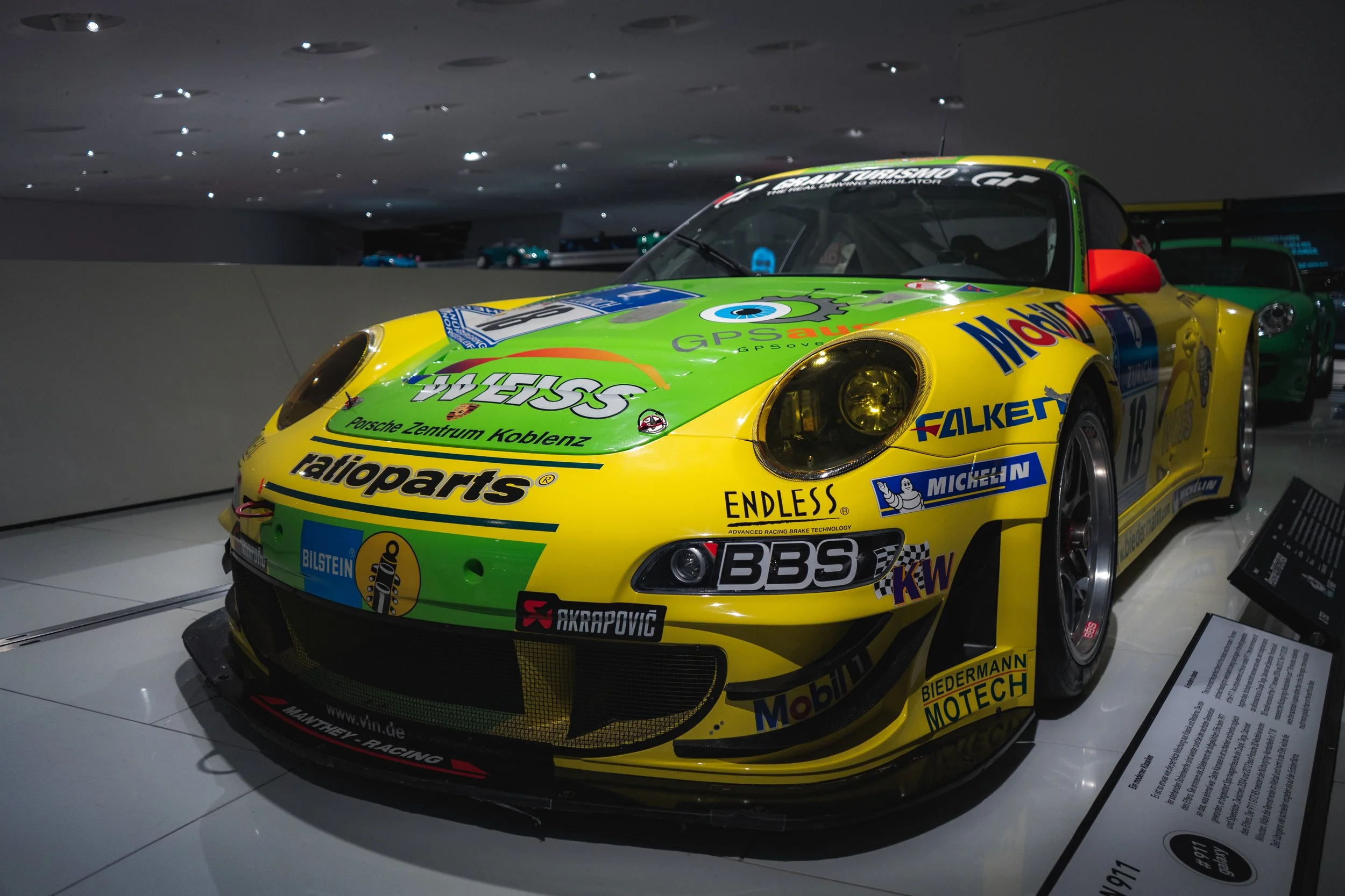 A brightly colored green and yellow Porsche race car on display at a museum, featuring various sponsor logos and a sleek aerodynamic design.