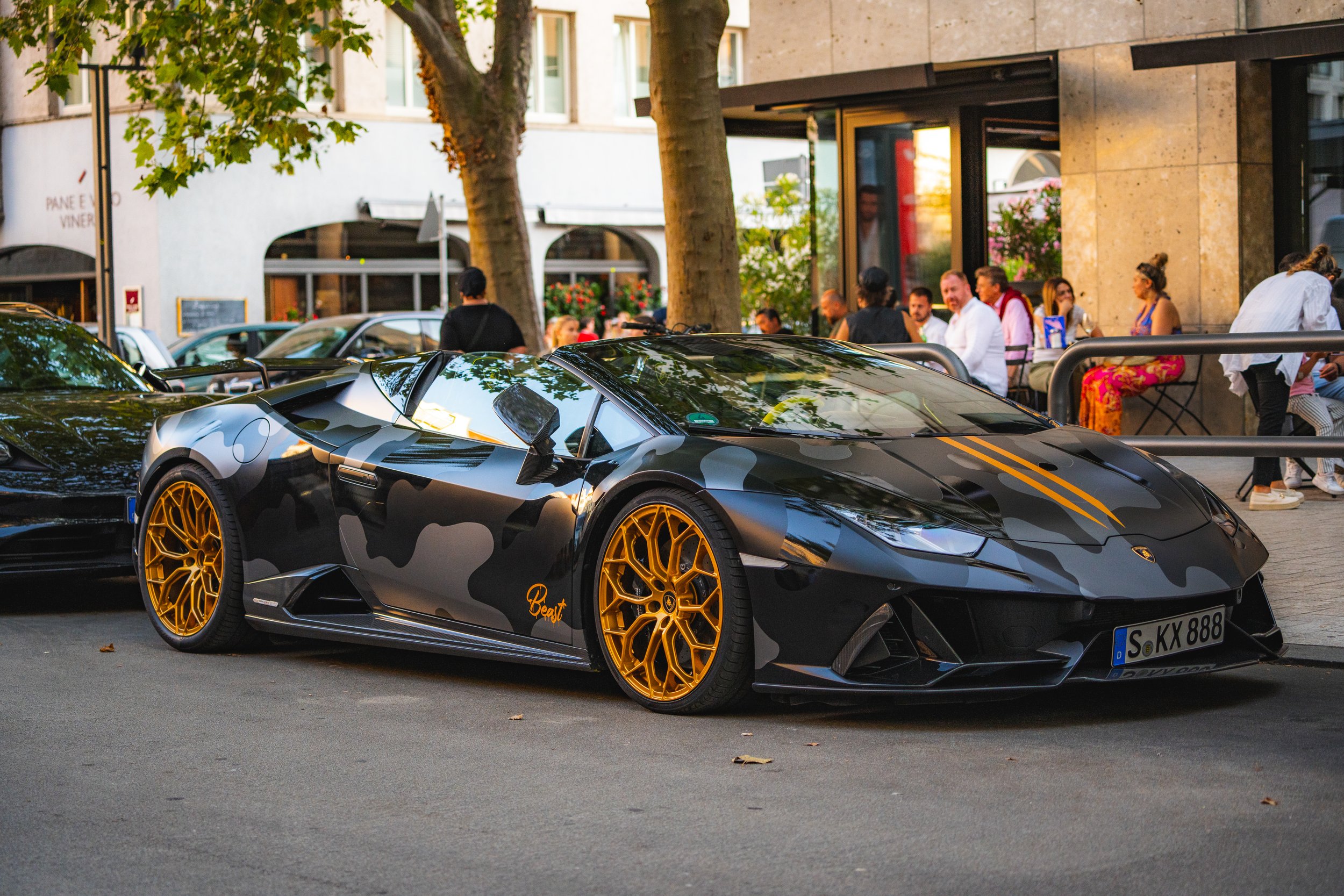 Black Lamborghini sports car with camouflage design and gold wheels parked on city street, with people sitting outside a restaurant or cafe in the background.