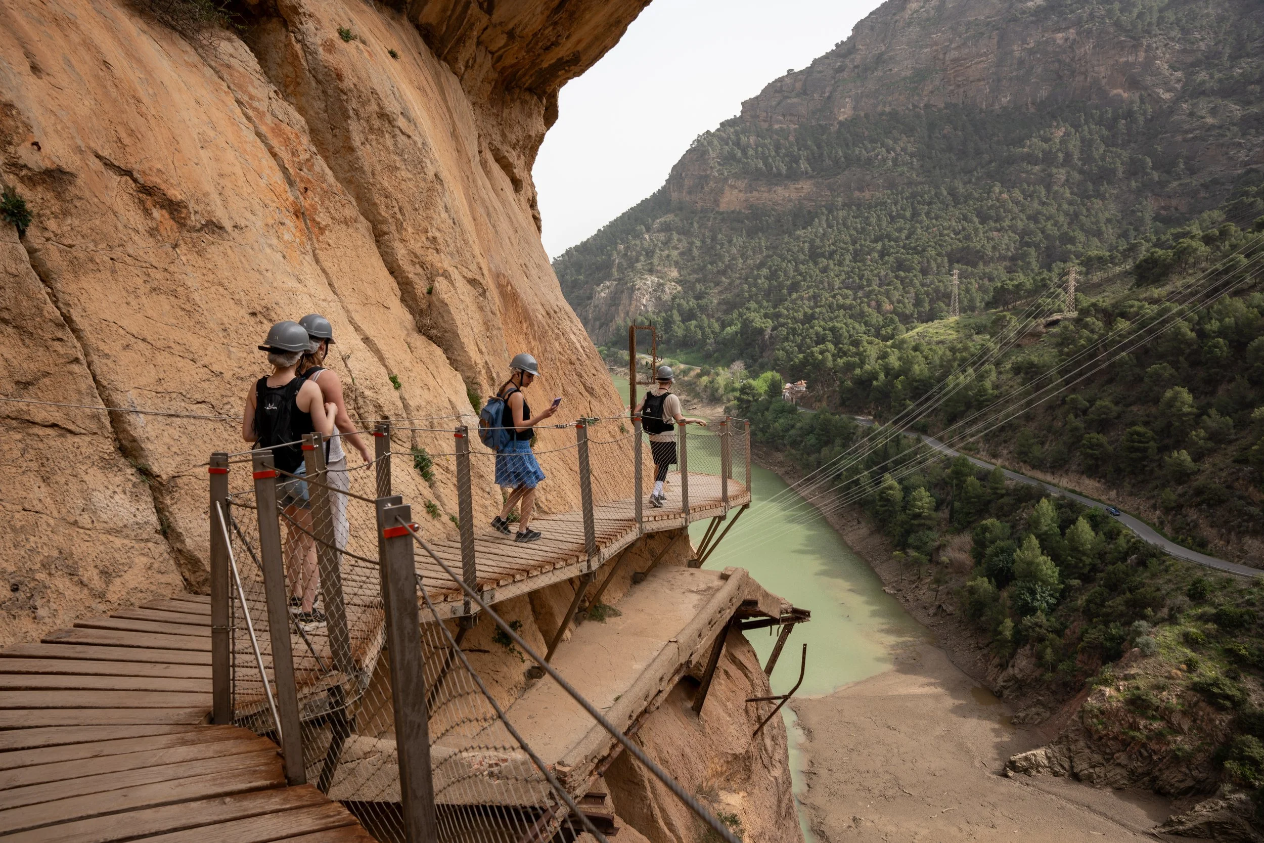 Tourists wearing helmets walking along a narrow suspended wooden walkway on a mountainside with a river below.