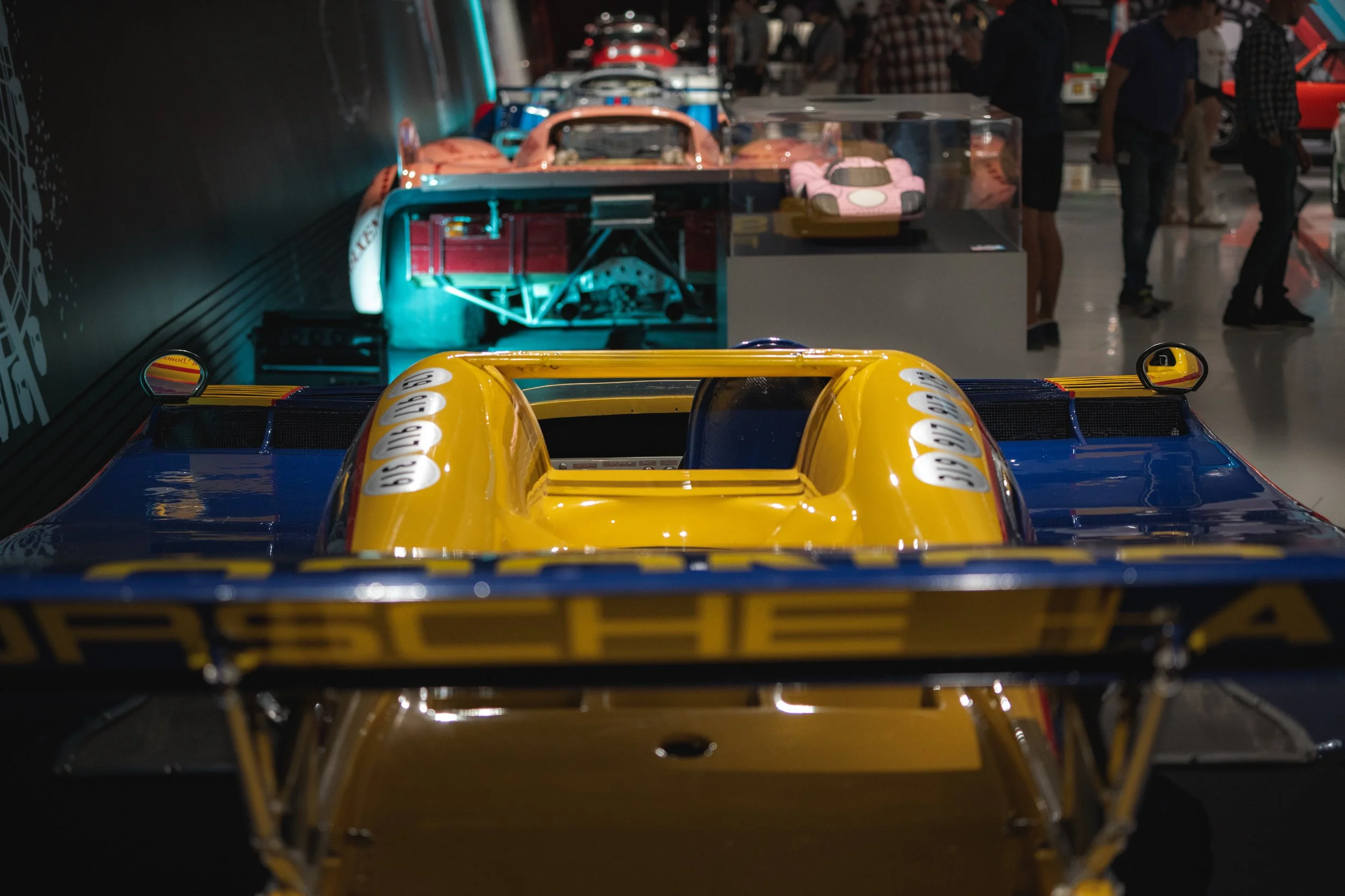 Close-up of the front of a vintage race car with a yellow and blue paint job, displayed at an exhibition with collectible cars in the background.