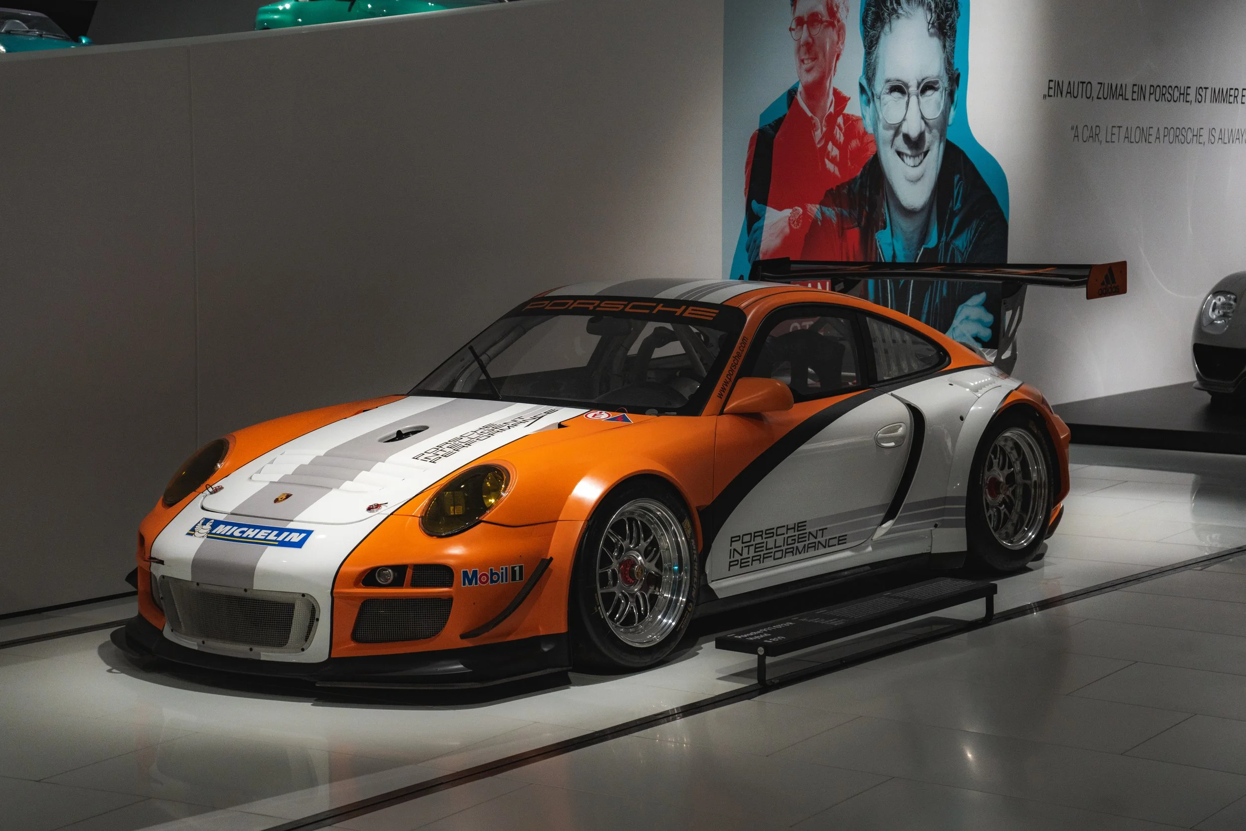 A Porsche race car on display, painted in orange, white, and black with sponsor decals, including Michelin and Mobil 1, in a showroom or museum setting.