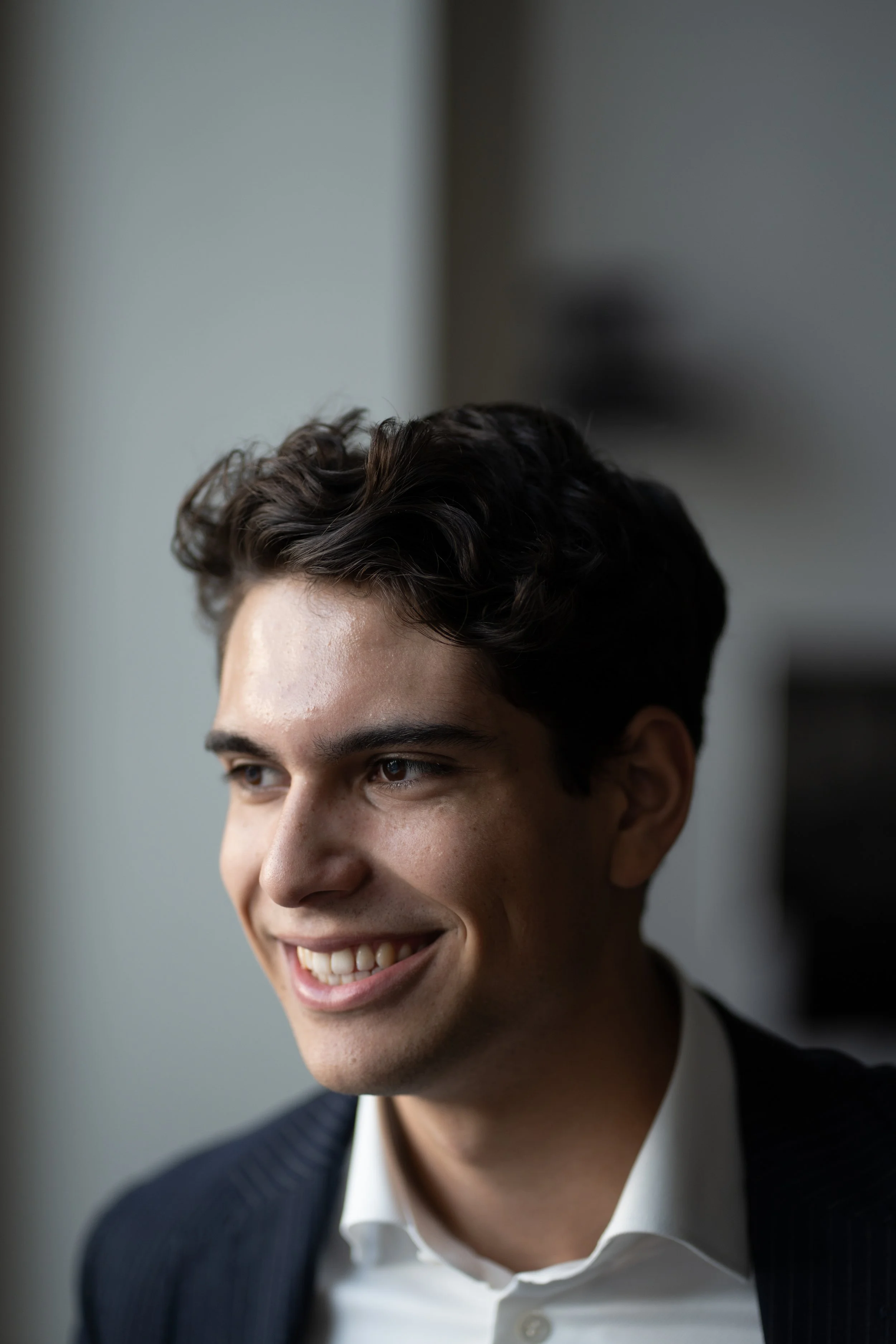 Young man in a suit smiling and looking to the side, indoors with natural light.