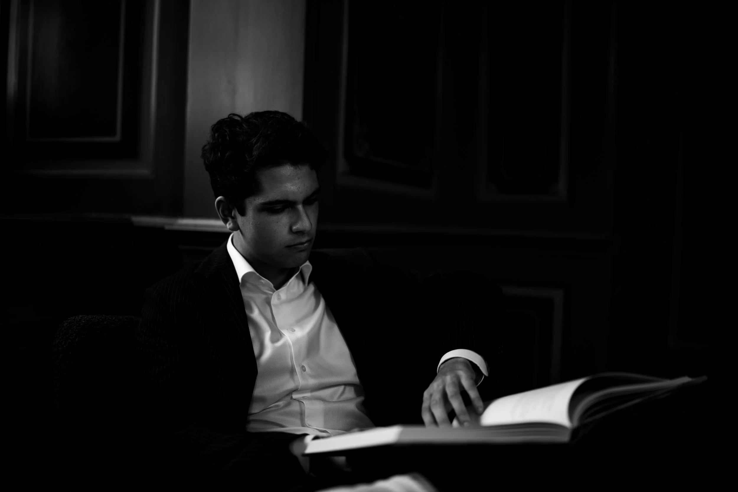 A man in a suit and white shirt reading a book in a dimly lit room with dark paneled walls.