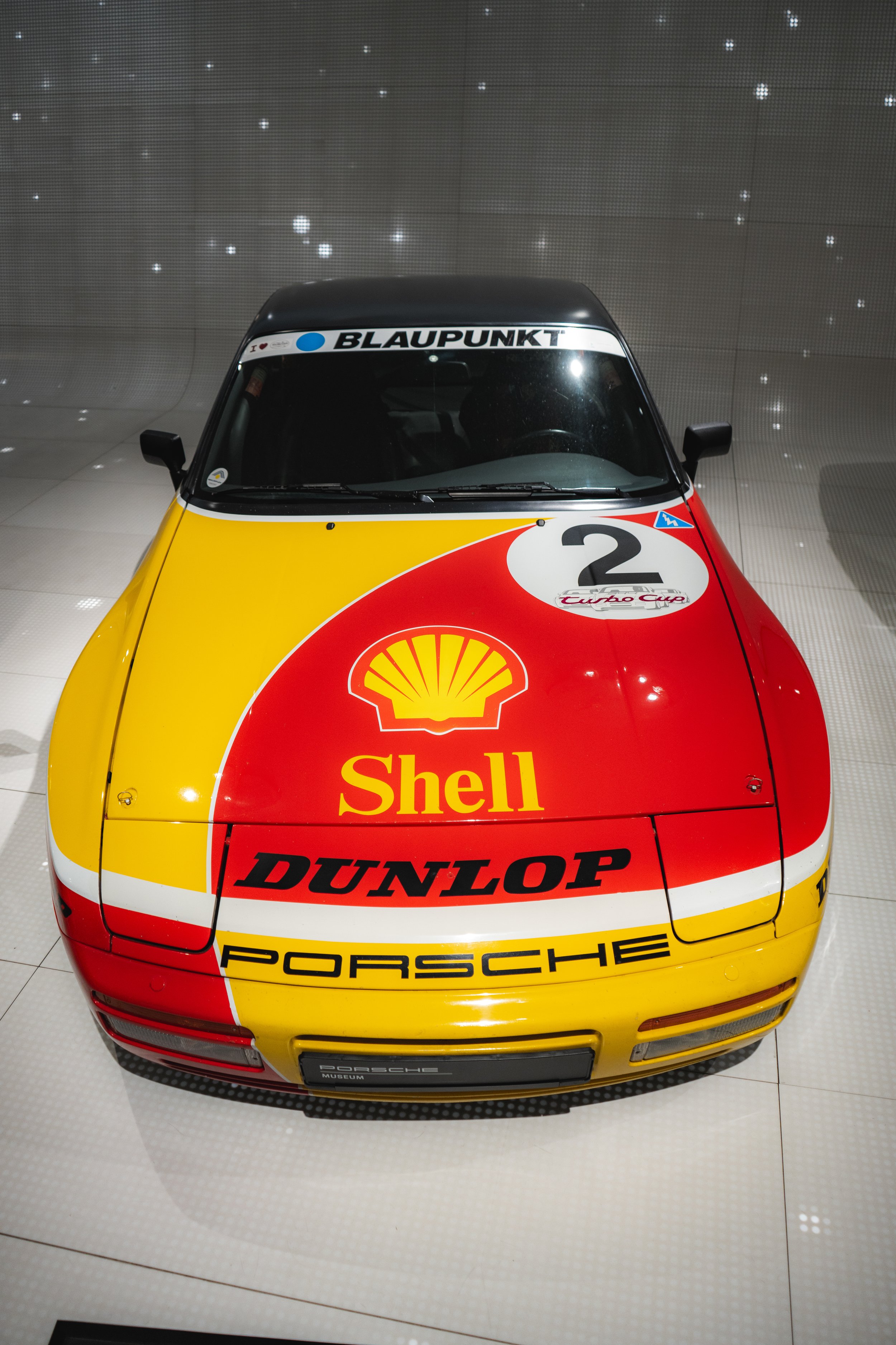 Front view of a vintage Porsche race car with yellow, red, and white Shell, Dunlop, and Porsche branding, displayed in a museum.