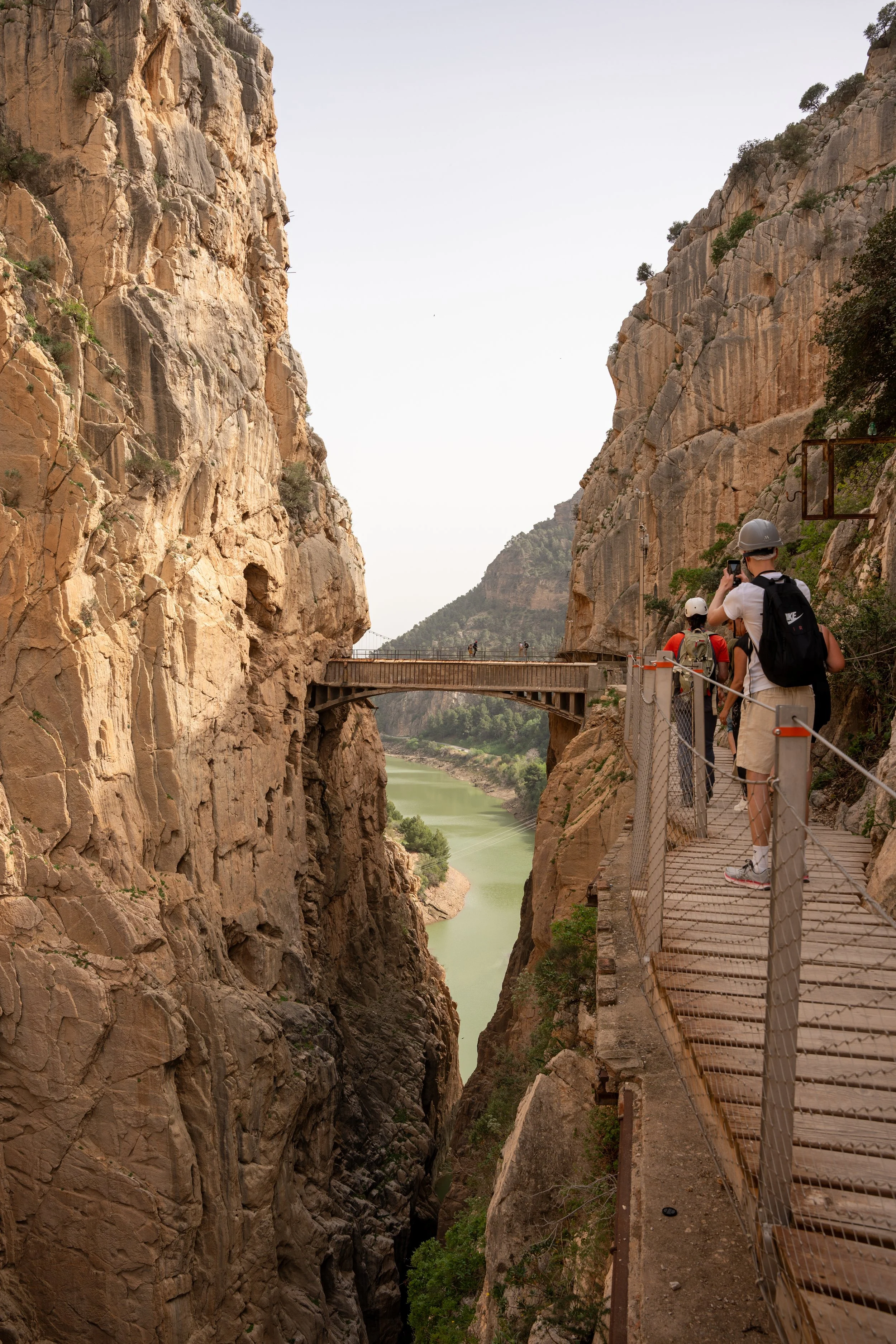 Tourists walk along a narrow suspended walkway with a metal railing, surrounded by tall rocky cliffs, overlooking a green river in a canyon.