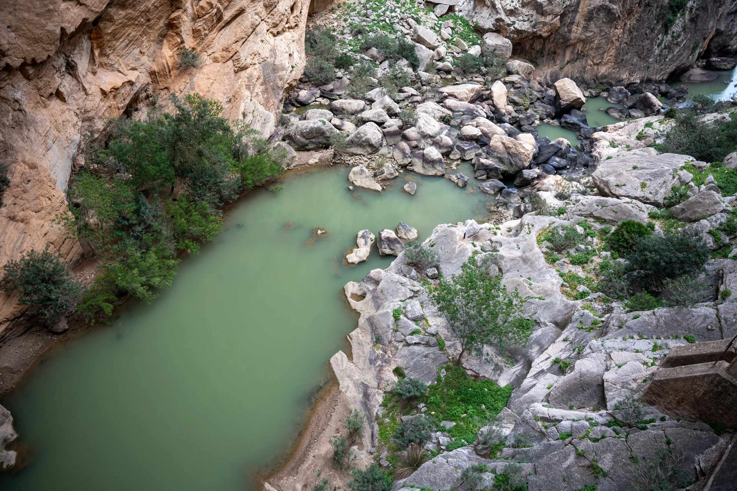 A high-angle view of a rocky canyon with a greenish river flowing through it, surrounded by cliffs and sparse green vegetation.