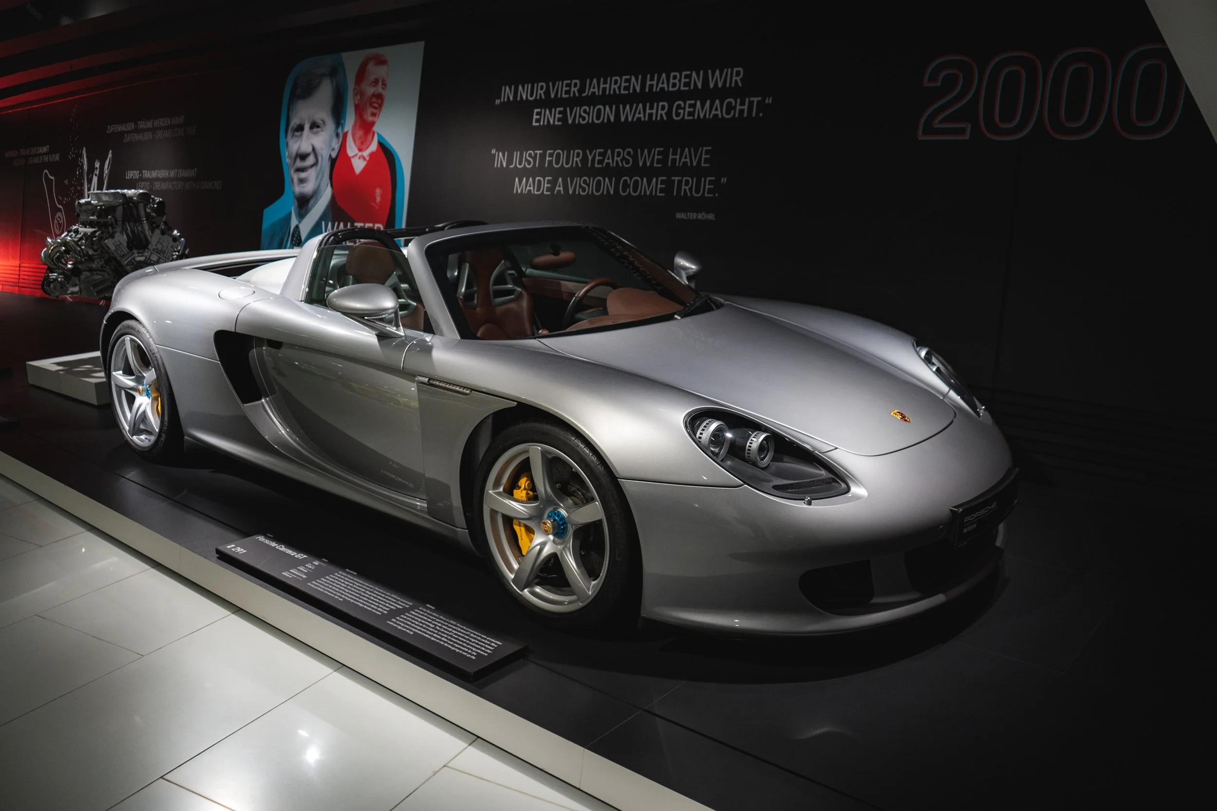A silver Porsche supercar displayed on a black platform in a showroom with a dark background and informational text, including portraits and quotes.