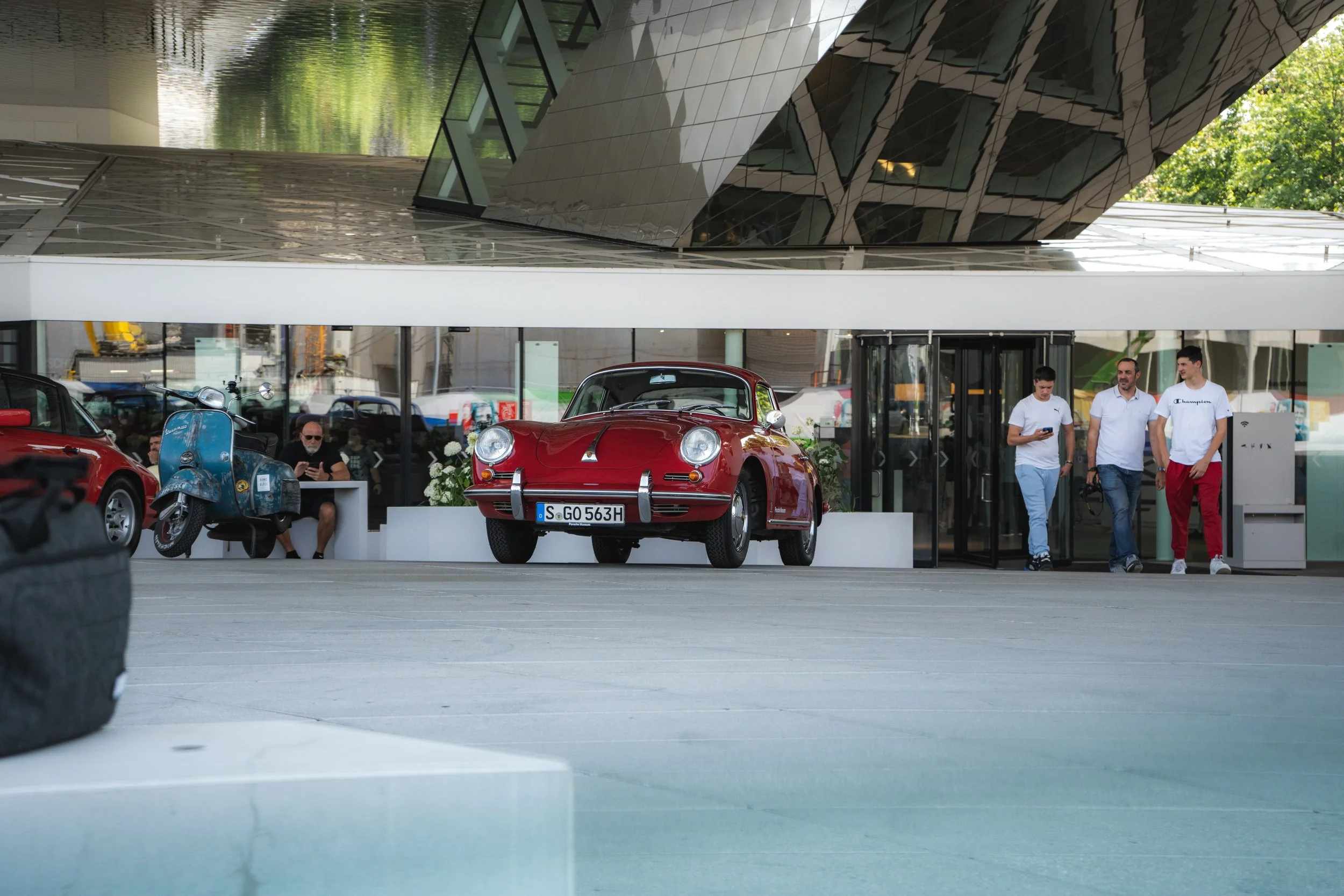 A red vintage Porsche sports car parked in front of a modern building with a glass façade. There are three men walking near the entrance, one of whom is wearing a white shirt and red pants. A man with a beard sits behind a small table, while a blue s