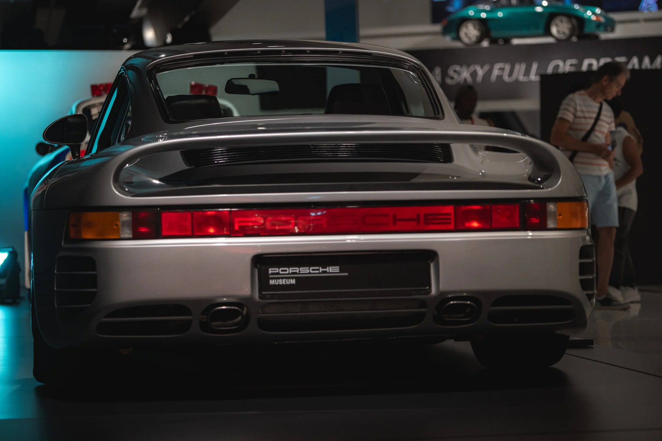 Rear view of a silver Porsche 911 sports car on display at the Porsche Museum, with a focus on the car's tail lights, exhaust, and the Porsche badge.