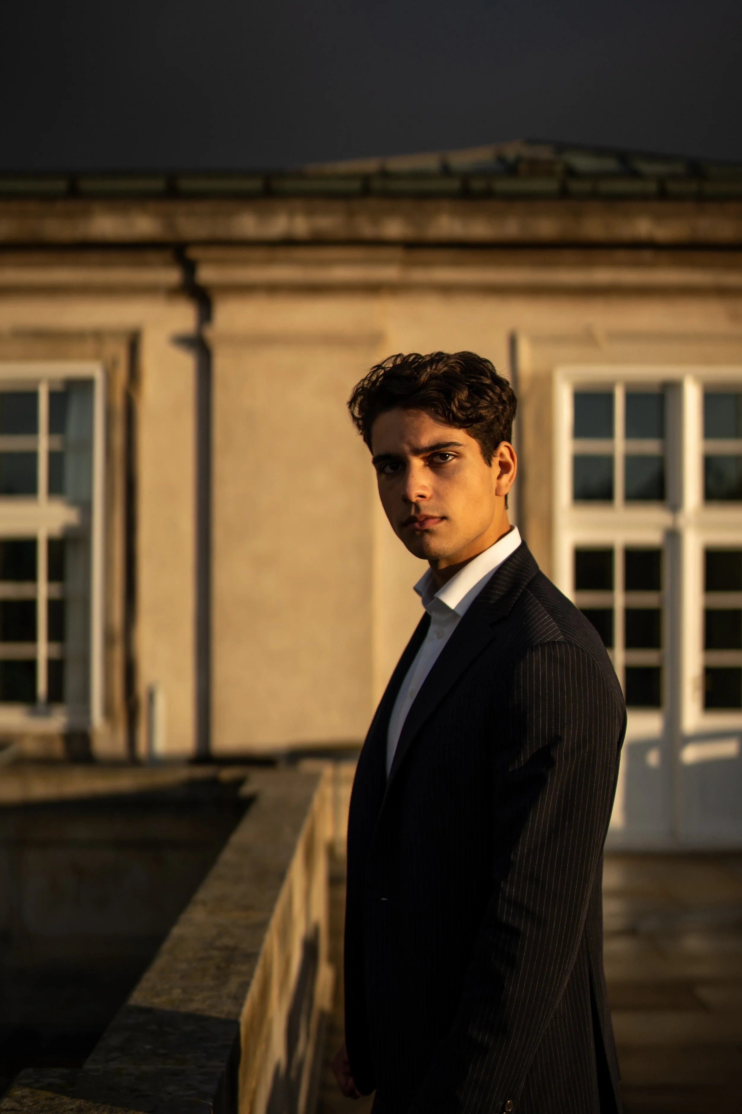 A young man dressed in a black suit and white shirt stands on a balcony at sunset, with a building with large windows in the background.