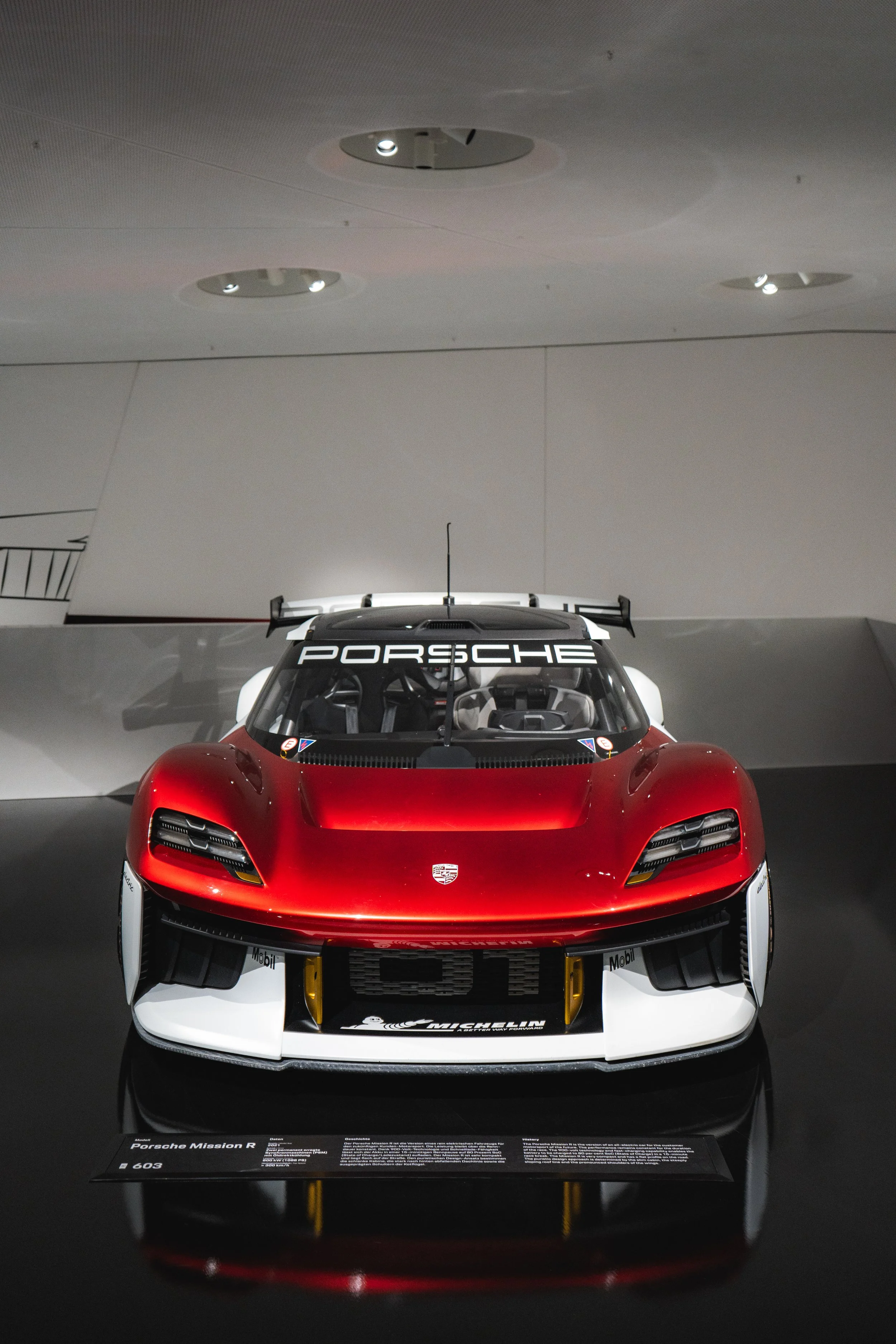 A red Porsche Mission R race car on display in a museum with a black informational plaque in front.