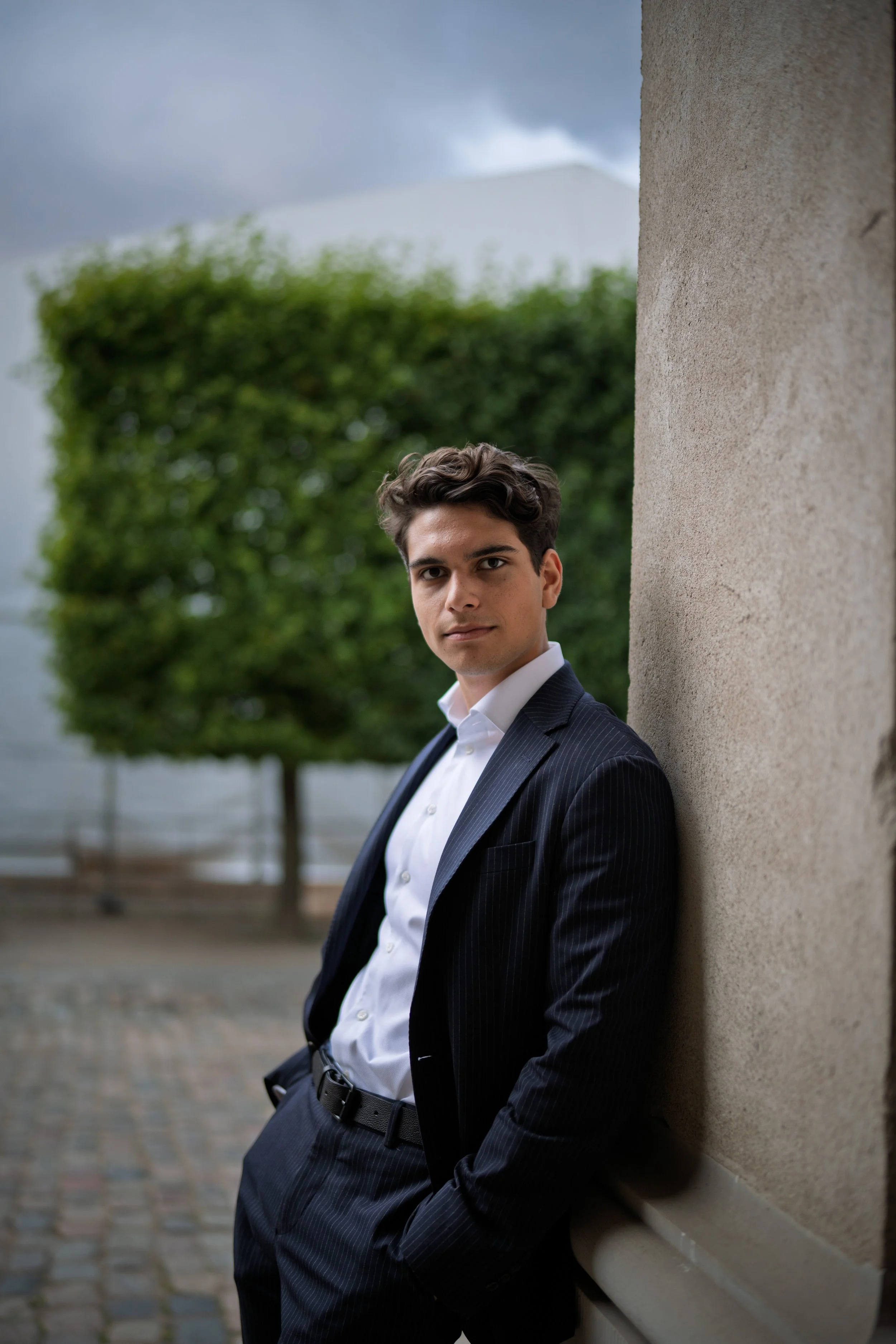 A young man in a dark suit and white shirt leaning against a textured wall outdoors, with a blurred tree and cloudy sky in the background.