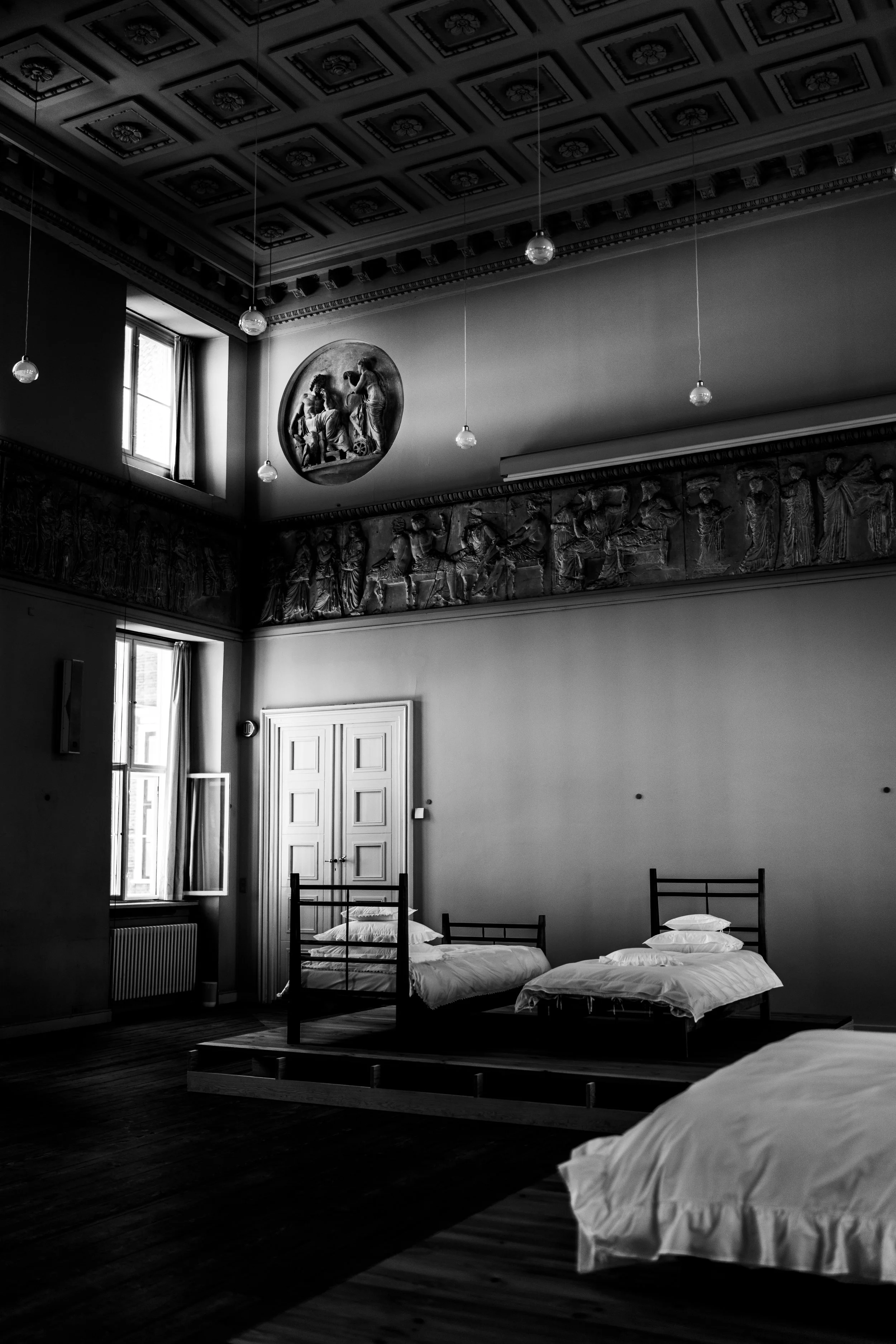 Black and white photo of an interior room with high ceiling, decorated with framed ceiling panels, hanging lights, windows, and classical wall reliefs. Three beds with white bedding are visible, one partially in view.
