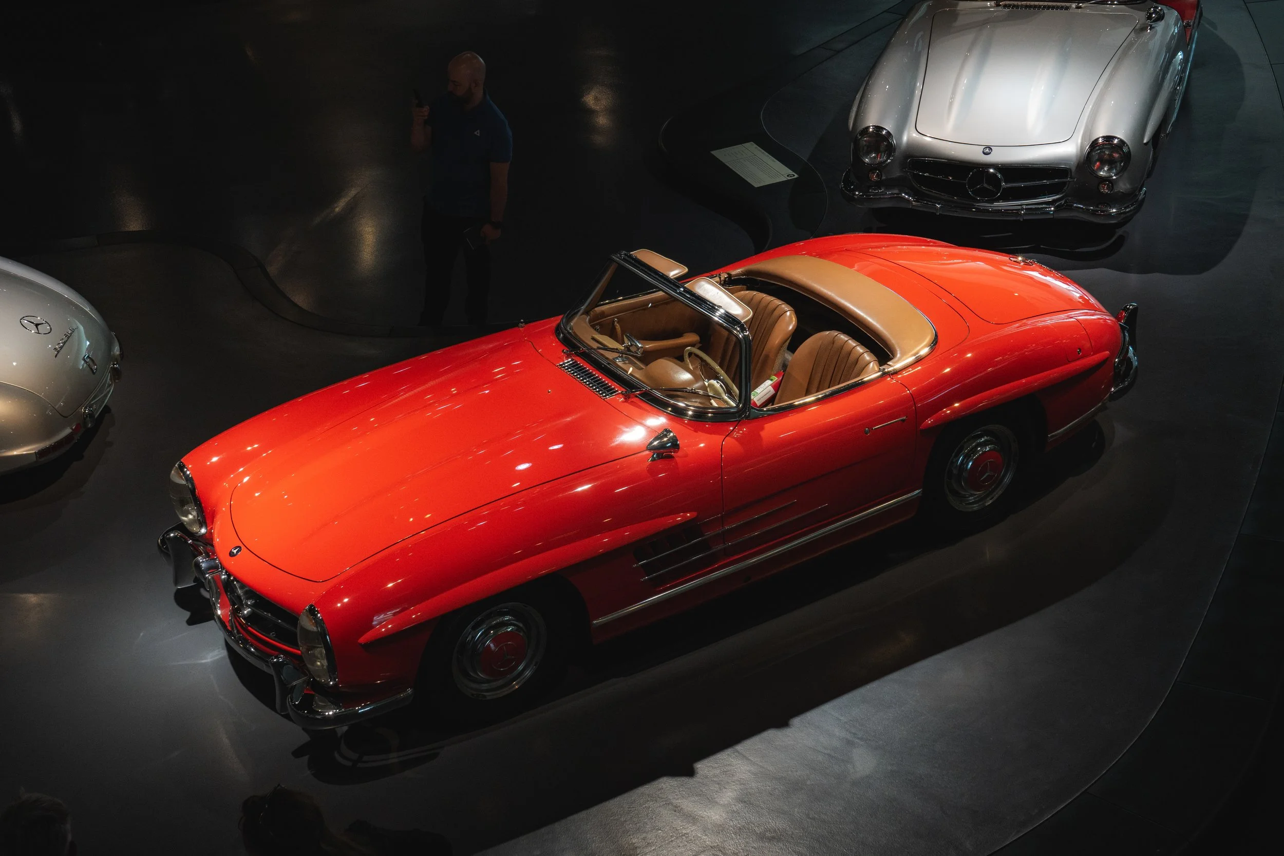 A classic red Mercedes-Benz 300SL Roadster displayed in a museum, with tan interior and other vintage cars around it.