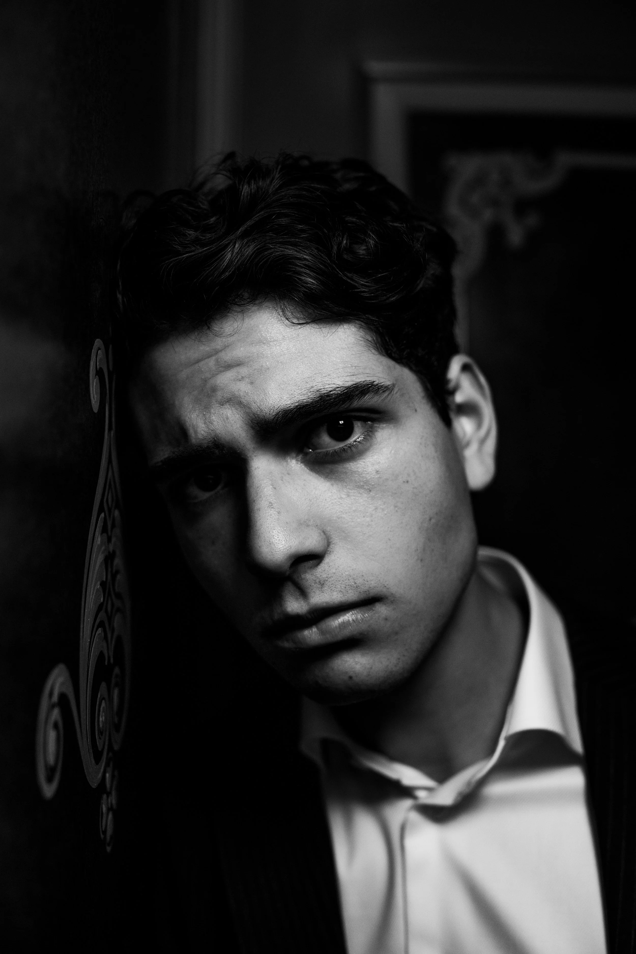 Black and white portrait of a young man with dark, curly hair, wearing a white shirt and dark blazer, leaning against a textured wall with artwork in the background.