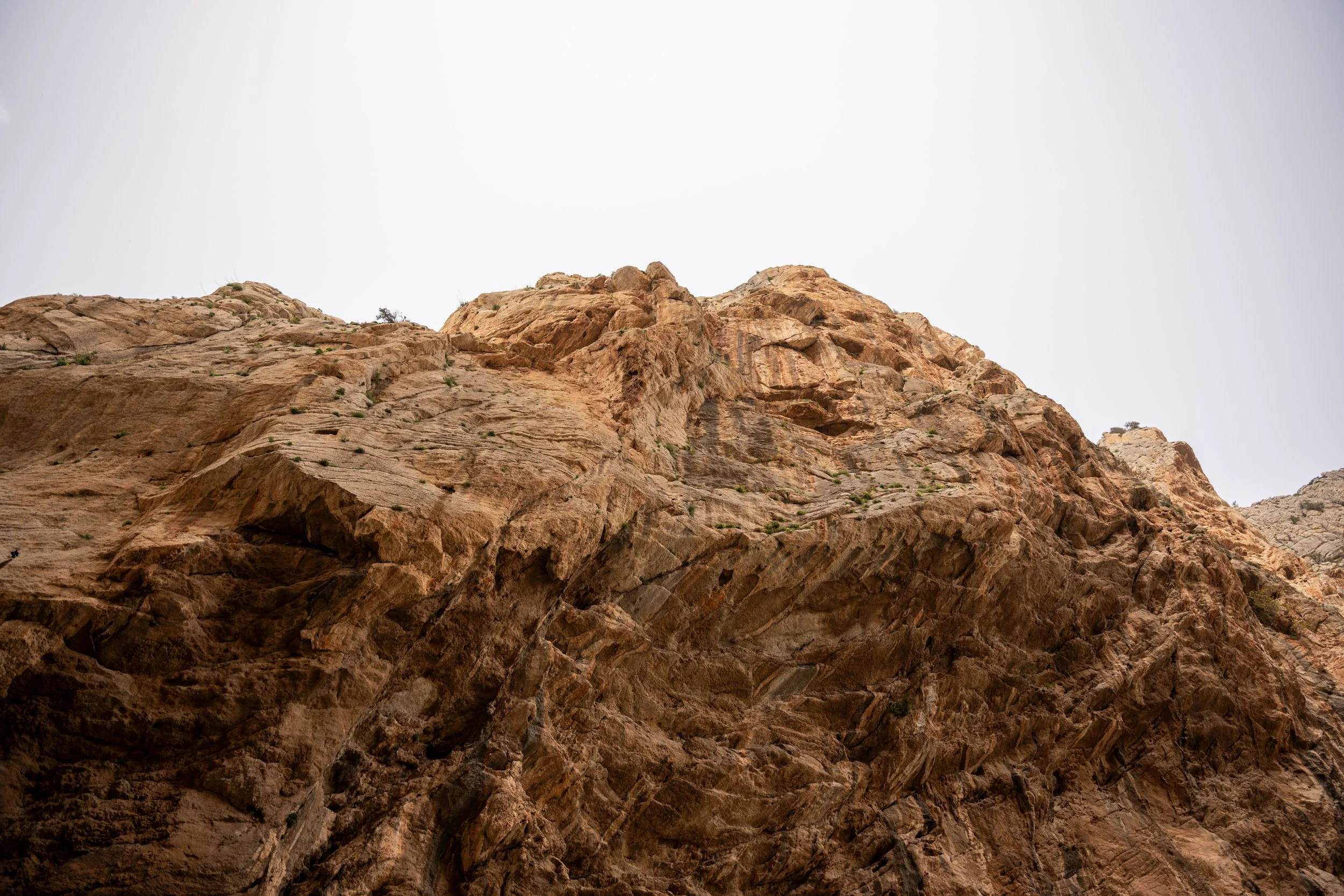 A close-up view of a rugged, reddish-brown rocky mountain with patches of sparse vegetation under a pale, overcast sky.