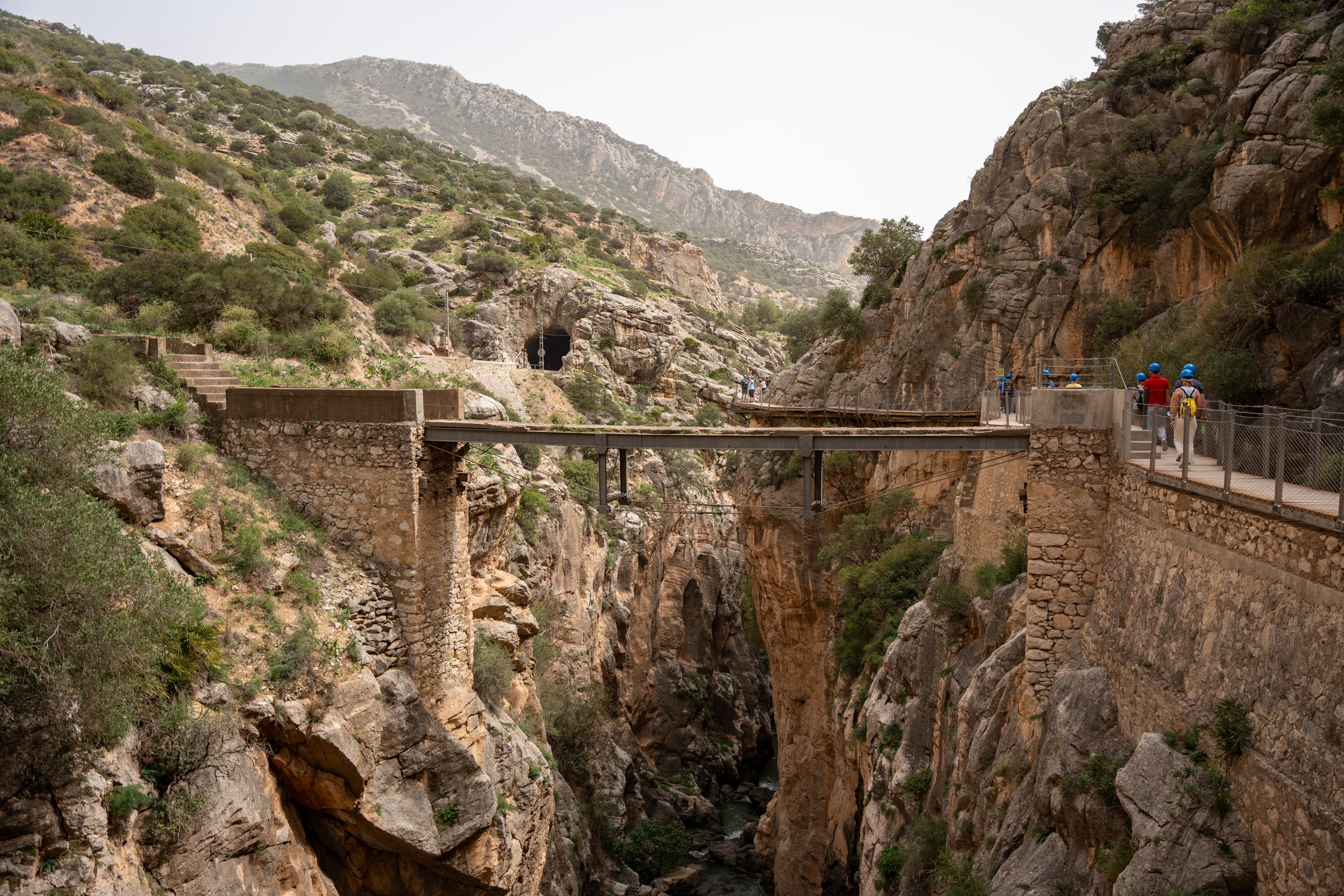 A group of tourists in blue helmets walking on a narrow mountain trail along a deep canyon with rocky cliffs, a tunnel in the mountain, and a small river at the bottom.