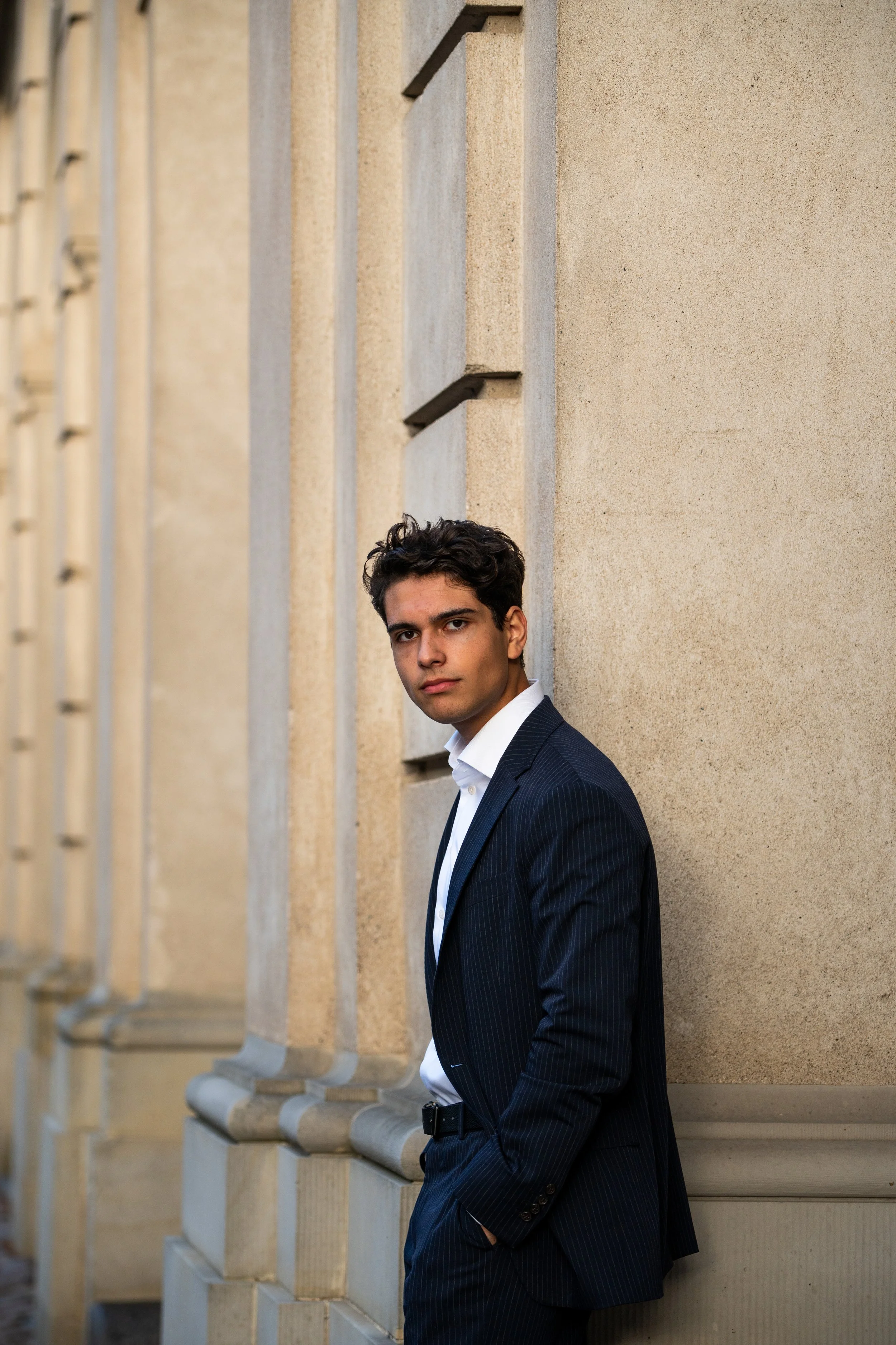 A young man in a dark pinstripe suit stands against a beige stone wall with architectural details, hands in pockets, looking at the camera.