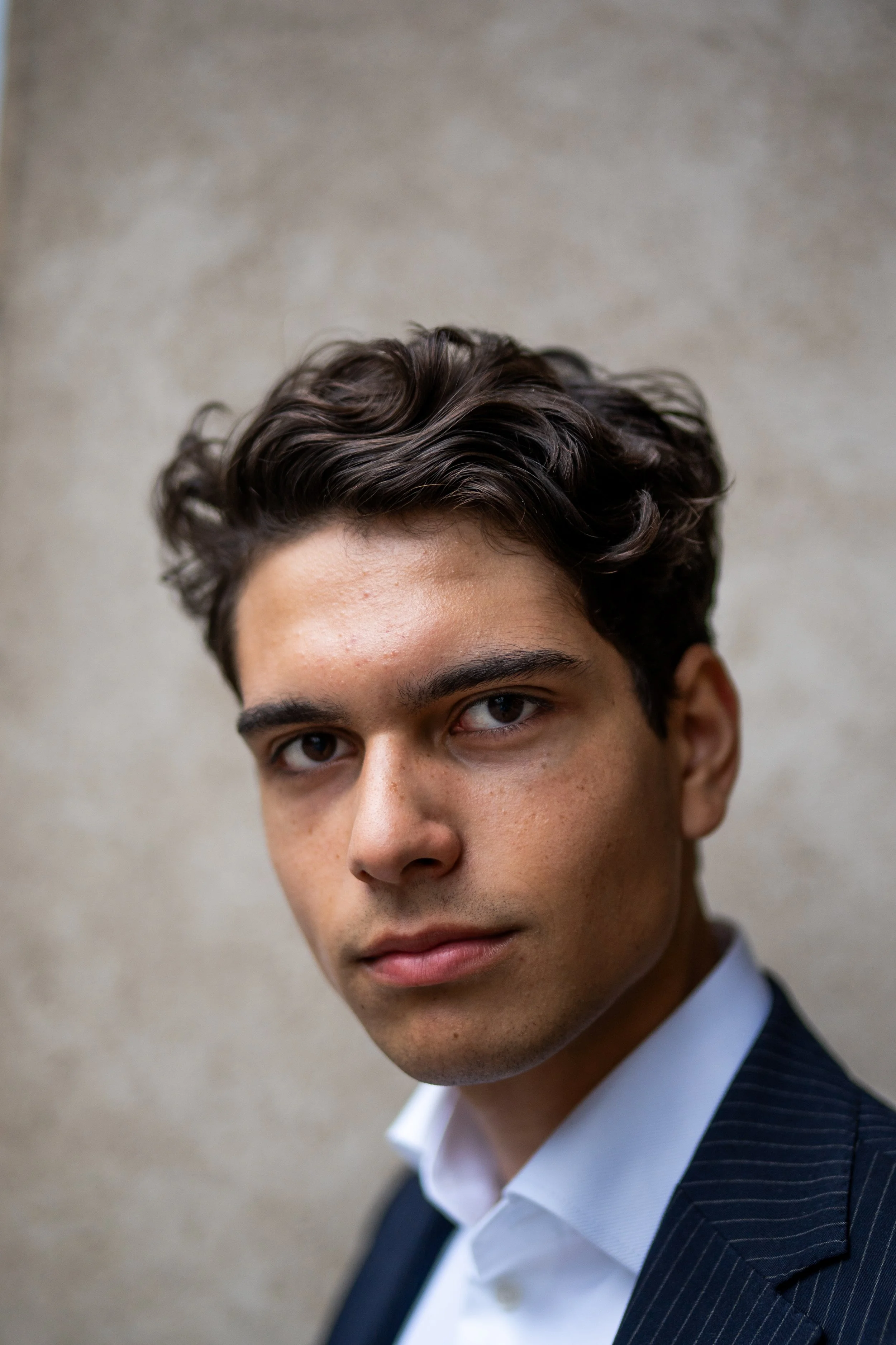 Close-up portrait of a young man with dark wavy hair, wearing a dark pinstripe suit and white shirt, standing against a beige wall.