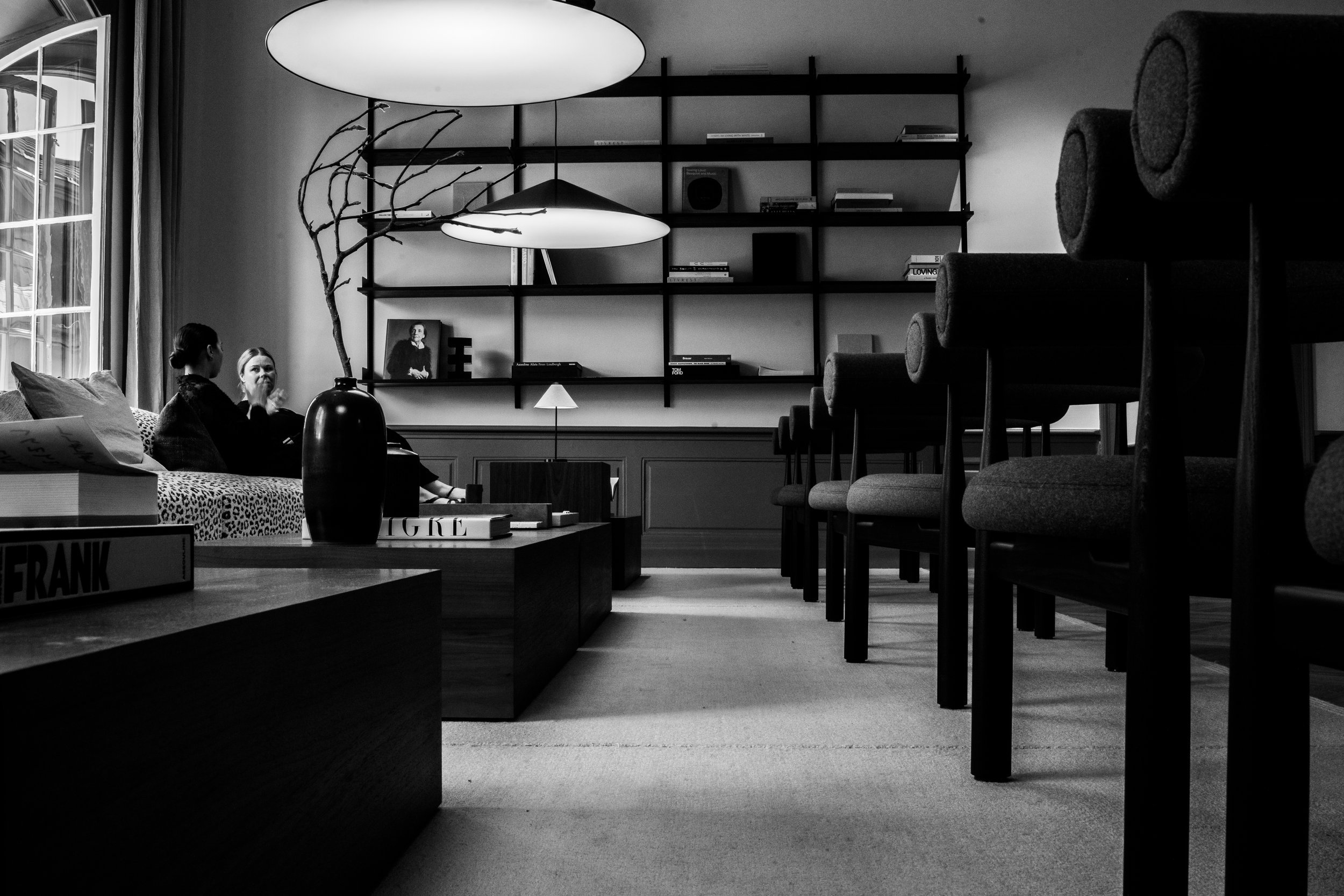 A black-and-white photo of a modern living room with two women sitting on a sofa near a window, facing each other, with a shelf and decor behind them, and multiple chairs aligned along a wall on the right.