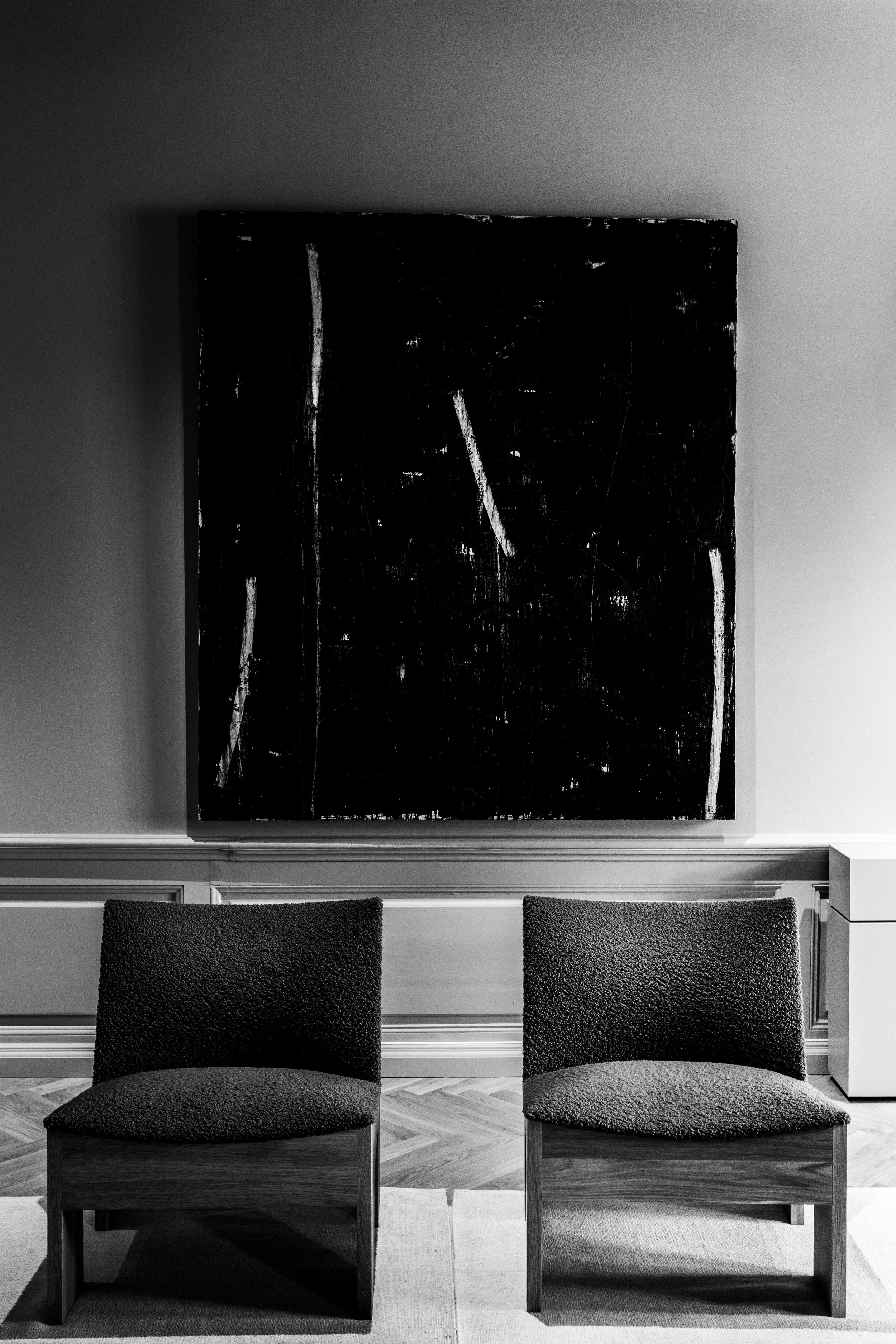 A black and white photograph of two chairs in front of a large abstract painting with white lines on a black background, in a room with wood flooring and decorative wall paneling.