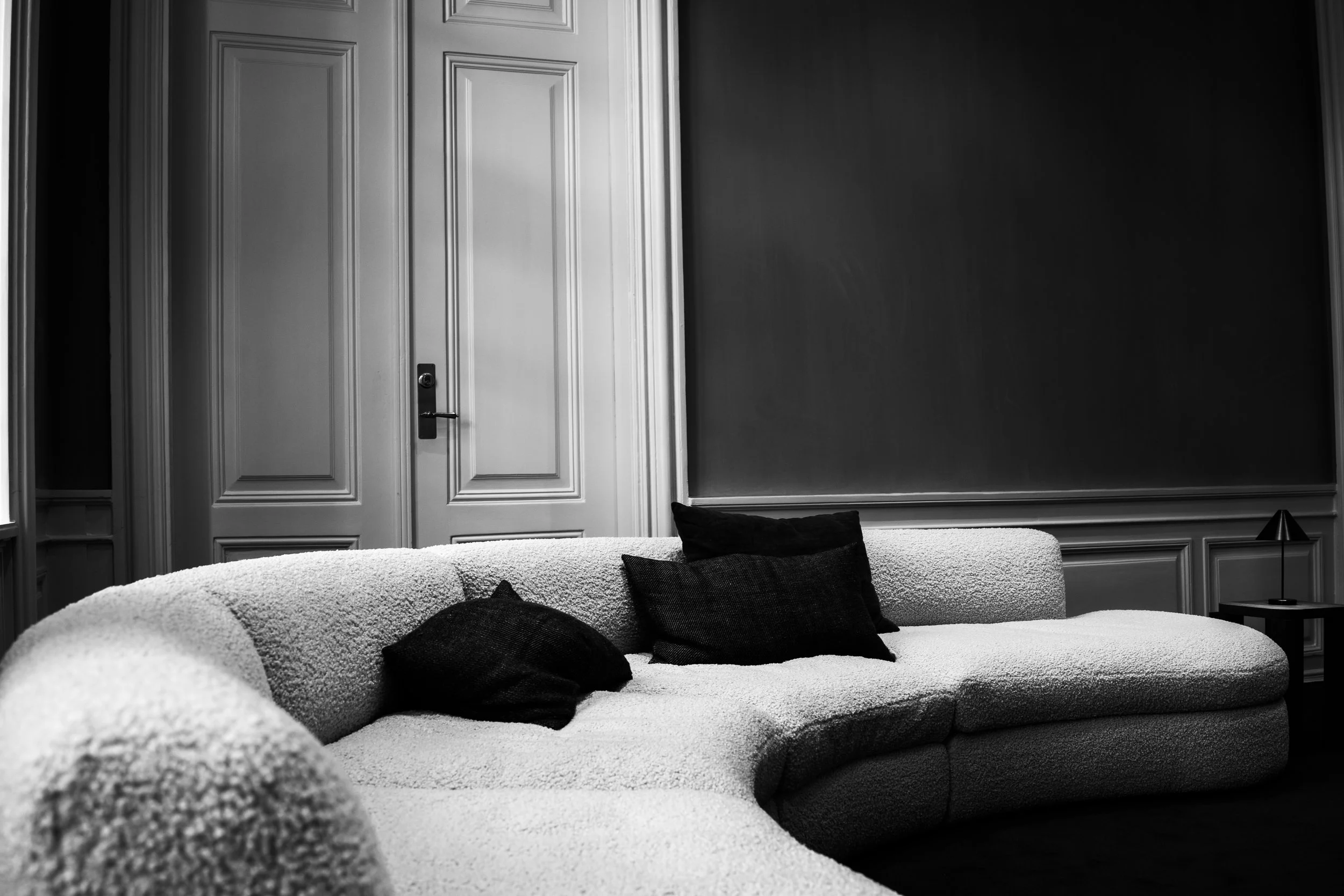 Black and white photo of a living room with a white textured sofa with three black pillows, a small black side table with a black lamp, a dark wall with paneling, and a closed double door in the background.