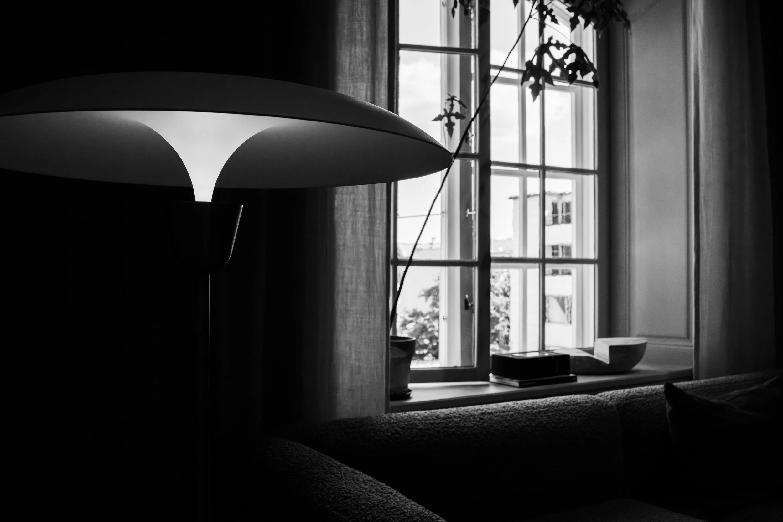 A black and white photo of a cozy interior space with a large window, a potted plant, books, and decorative bowls on the windowsill, and a modern lamp casting light.