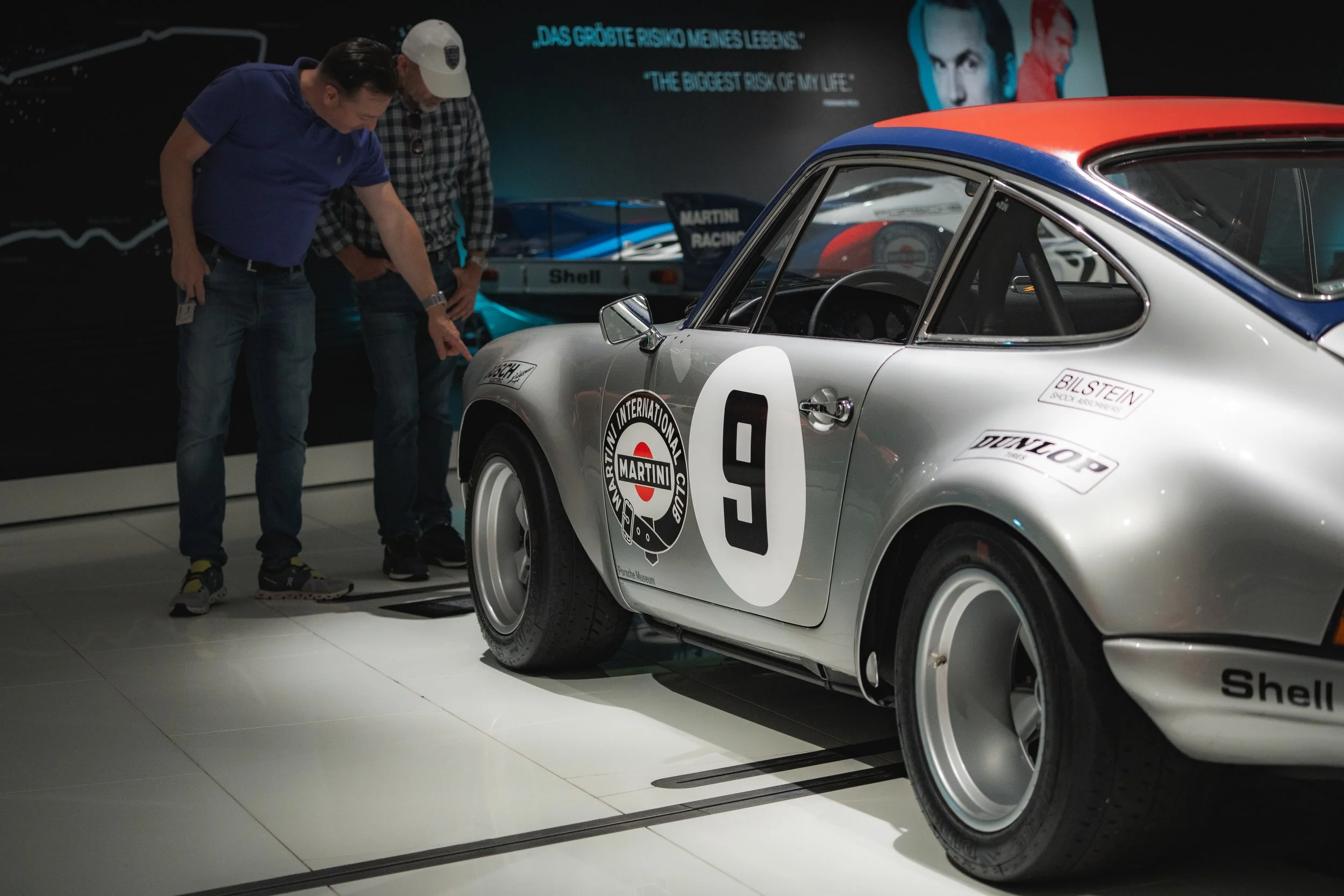 Two men examining a vintage white race car with the number 9 on its side at a museum or exhibit, with racing stickers and sponsors like Shell and Dunlop, in front of a black historical racing image.