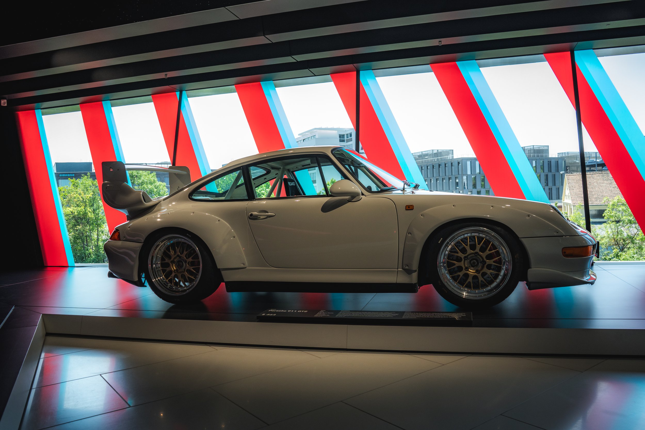 A classic white sports car displayed inside a modern building with large glass windows and colorful architectural design in the background.