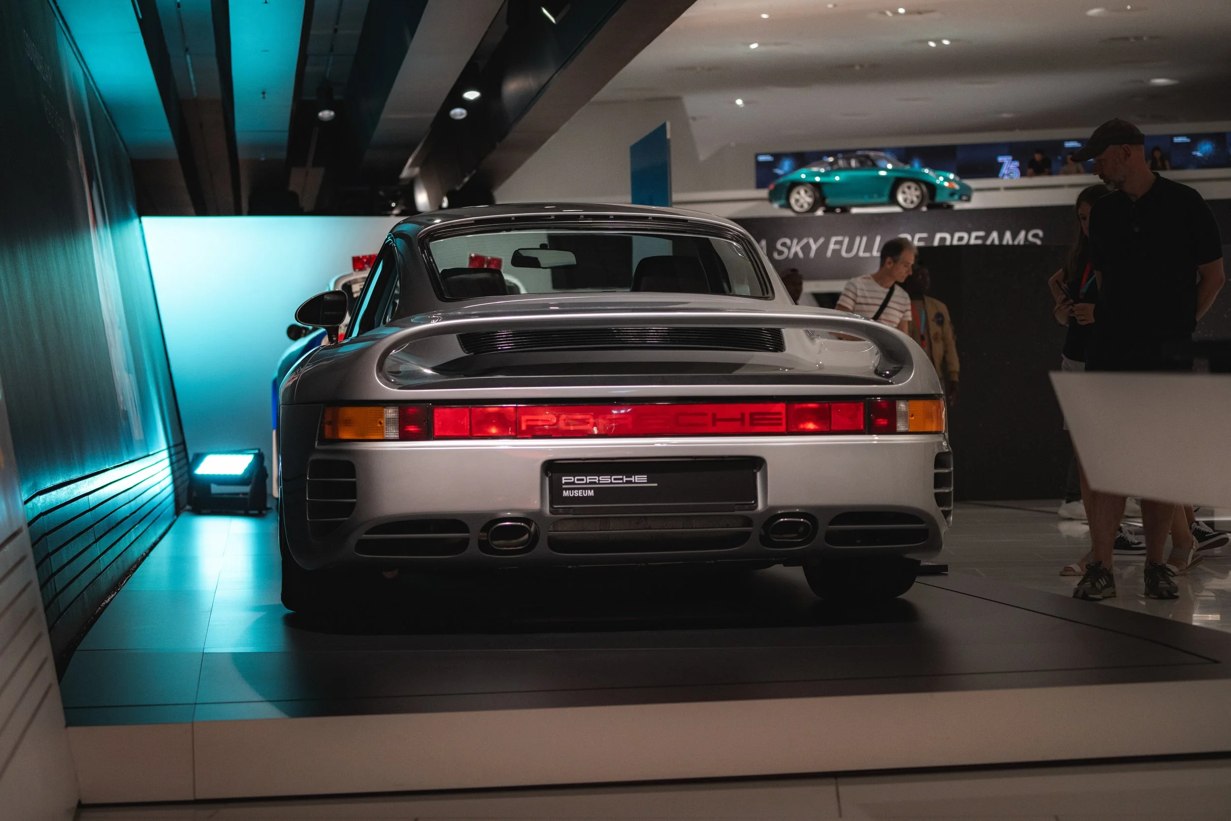 A silver Porsche 959 sports car on display at a museum, seen from the rear, with a distinctive tail light design and Porsche logo.