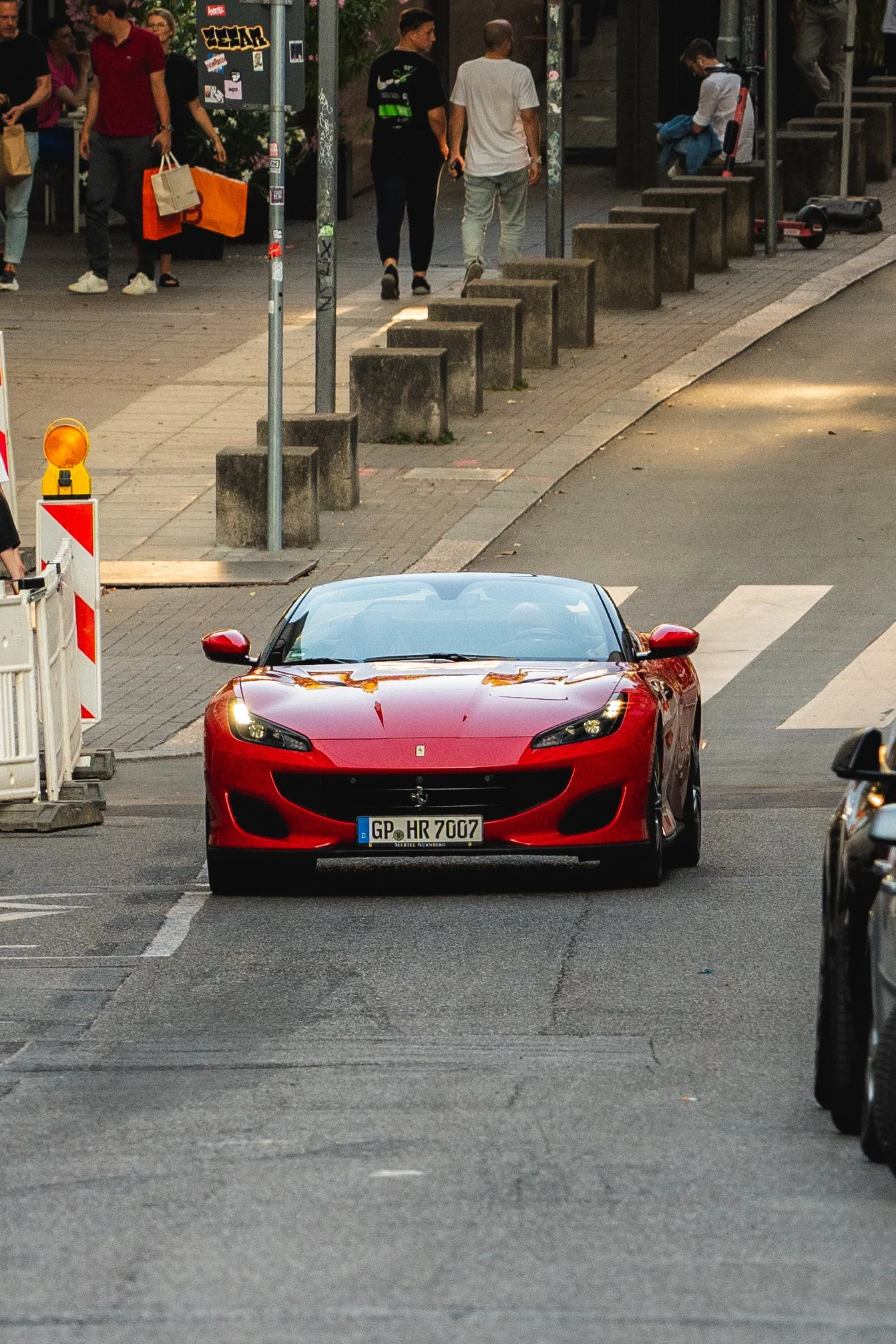 A red Ferrari sports car stopped in a city street crossing, with people walking on the sidewalk in the background.