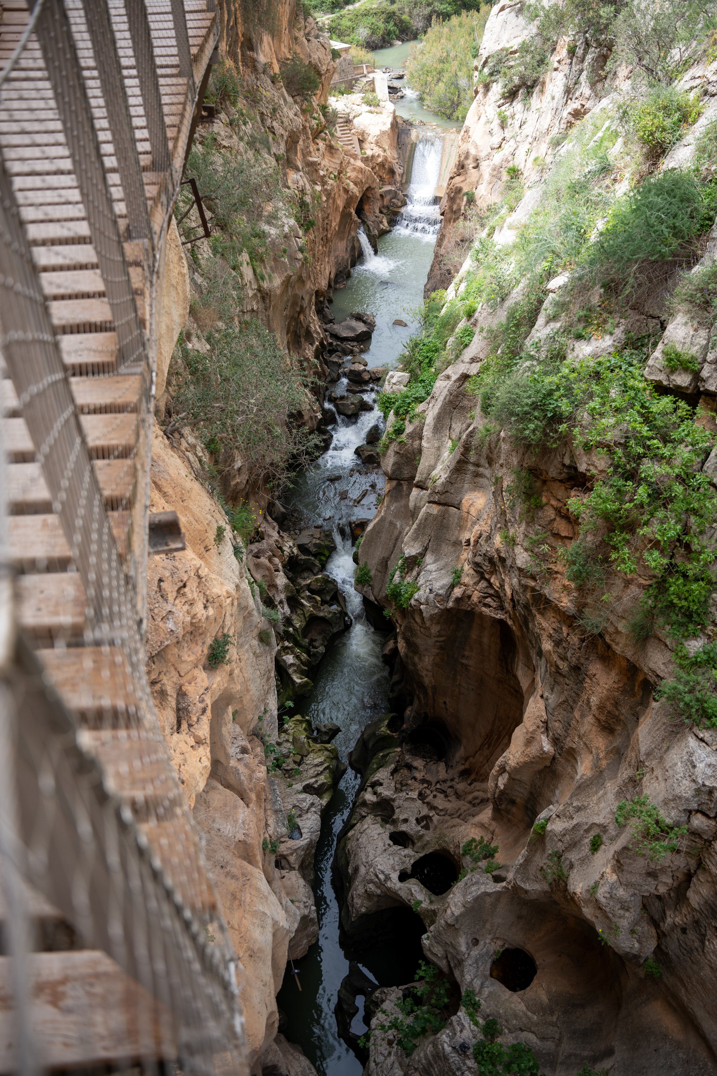 A narrow canyon with a flowing river and small waterfalls, viewed from a metal walkway on the left side, surrounded by rocks and green vegetation.