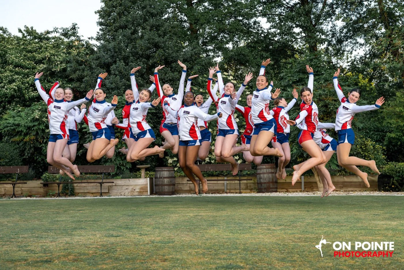 Group of cheerleaders in white, red, and blue uniforms jumping in unison on a grassy field with trees in the background.