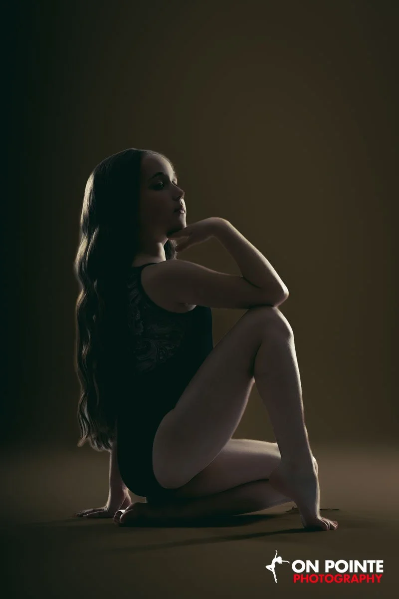 A woman with long, wavy hair sitting on the floor in a dark room, illuminated from the side, wearing a black lace dress, with one leg crossed over the other, her chin resting on her hand, in a contemplative pose.