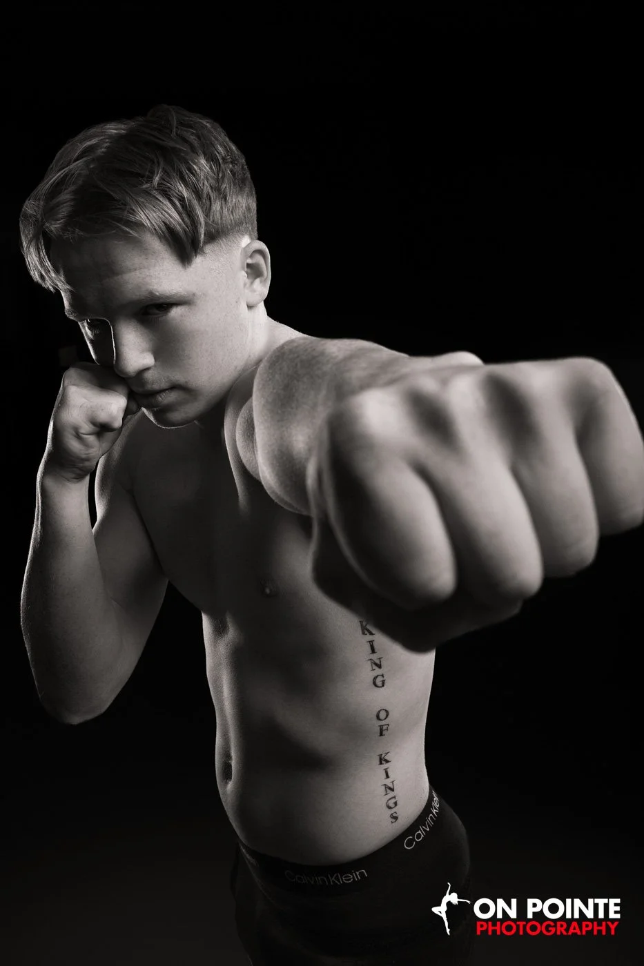 Black and white photo of a shirtless male boxer in a fighting stance, with "King of Kings" tattoo on his side and wearing branded underwear.